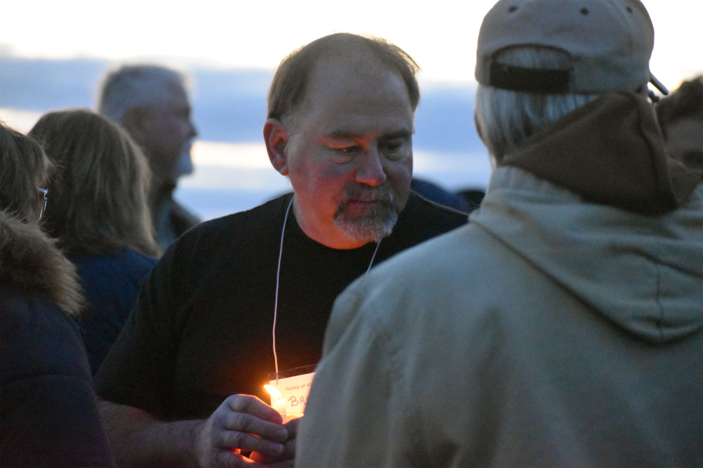 A man holds a lit candle and wears a name tag while talking to another person outdoors among a group of people at dusk.