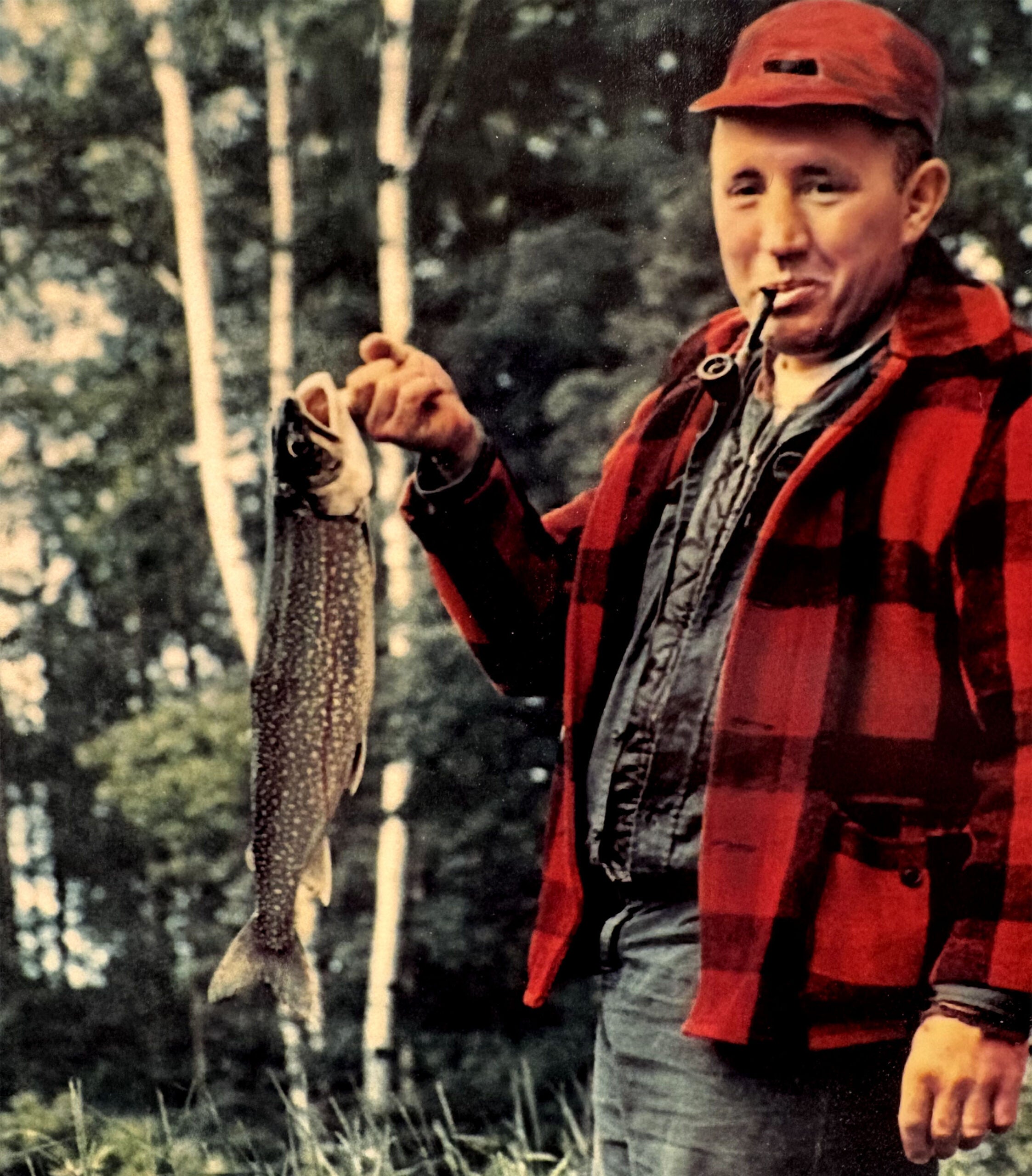 A man in a red plaid jacket and cap stands outdoors, holding a large fish in one hand and smoking a pipe. Trees are visible in the background.