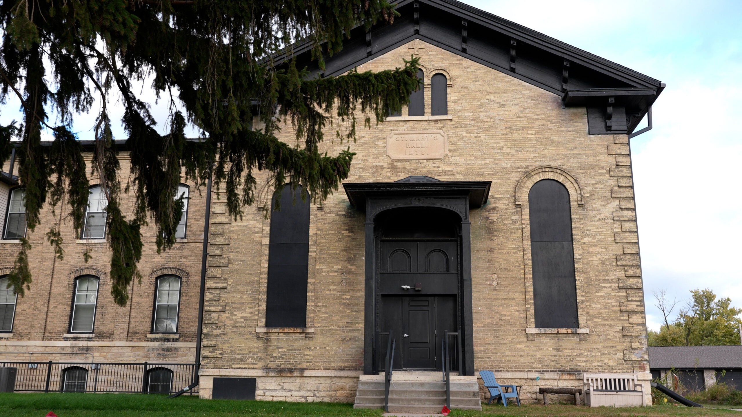 A large, old brick building with arched windows and a central entrance, partially obscured by tree branches in the foreground.
