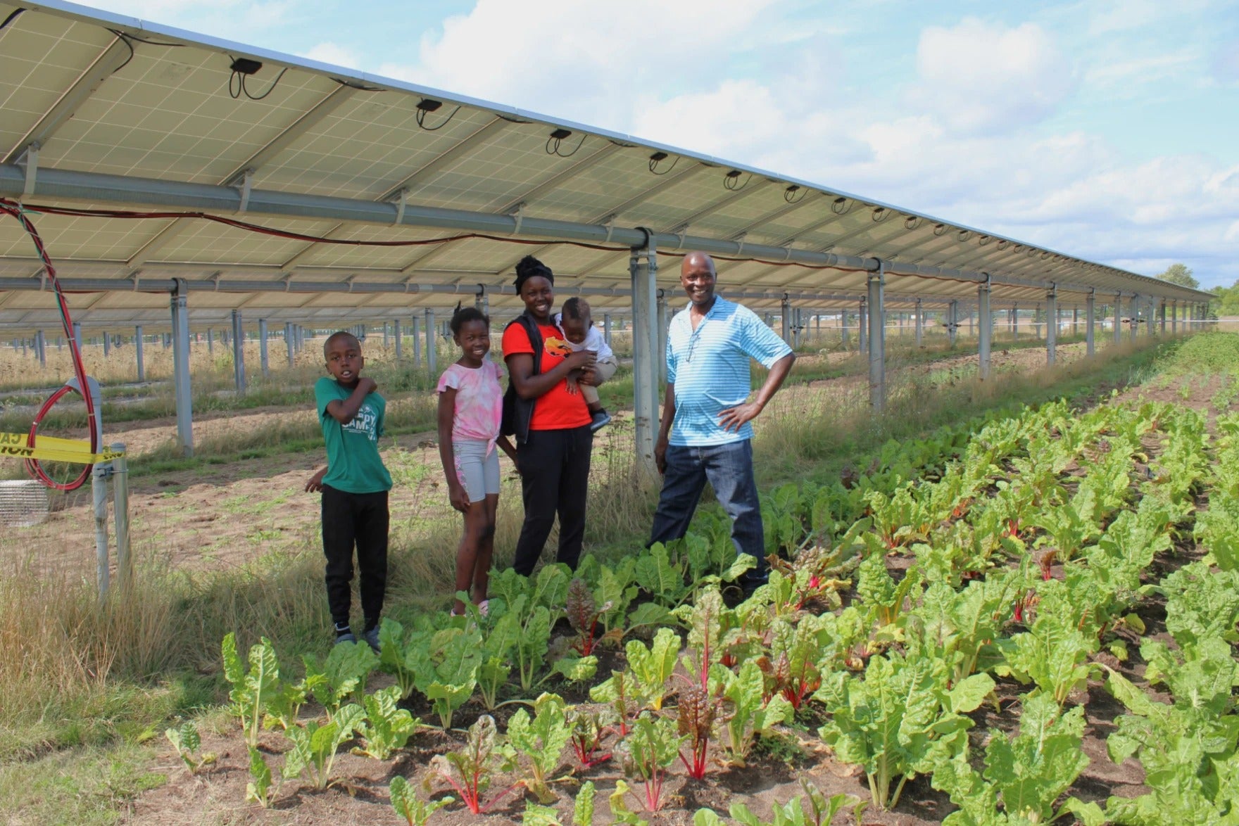 A family of five stands in a vegetable garden beneath solar panels on a sunny day.