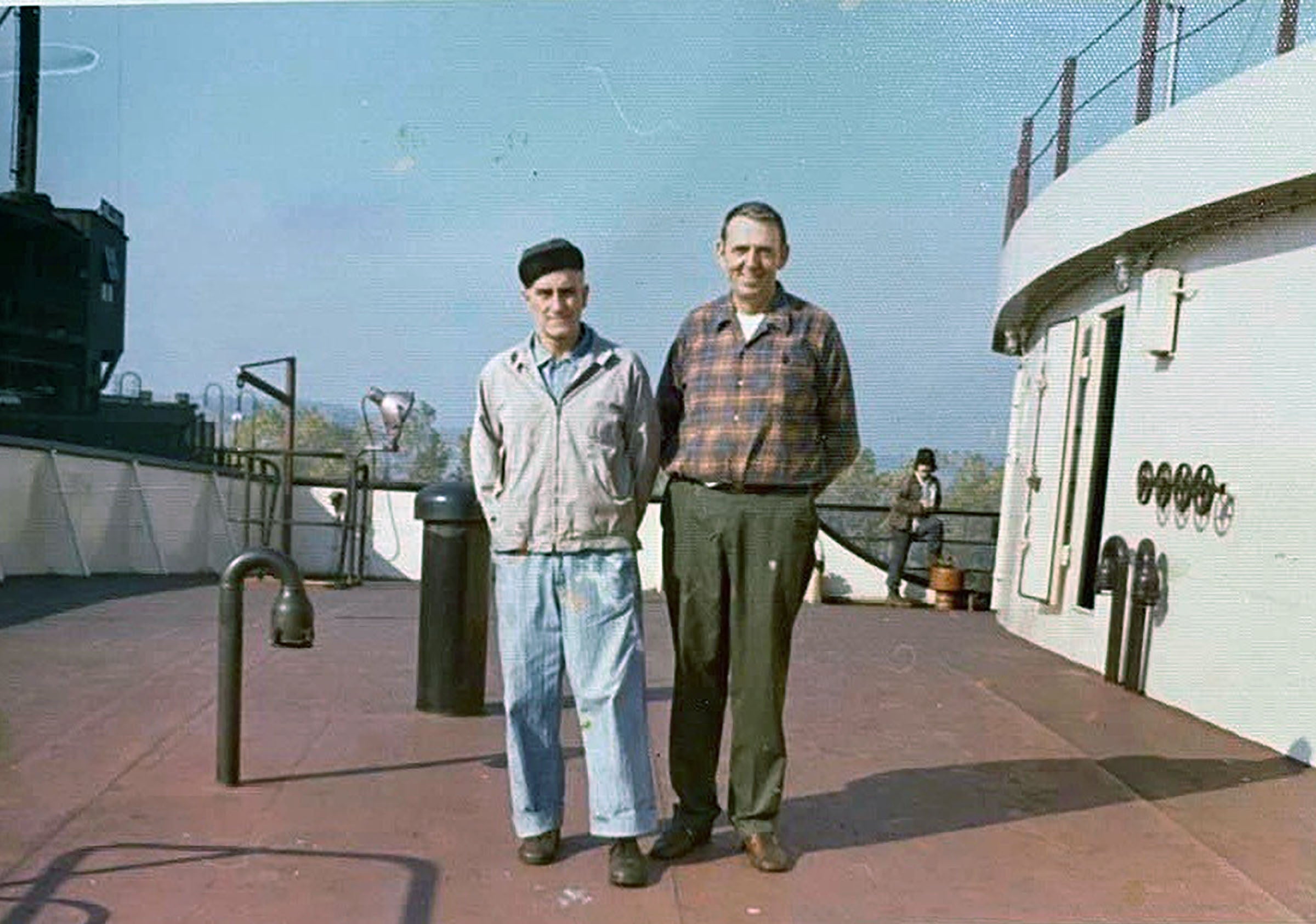 Two men standing on the deck of a ship, one in work clothes and a cap, the other in a plaid shirt and slacks, with ship equipment visible in the background.