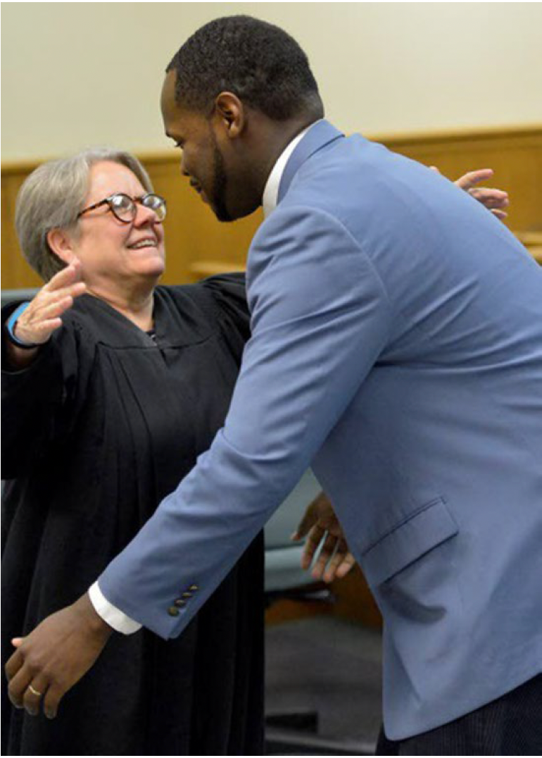 A judge in black robes smiles and prepares to hug a man in a light blue suit inside a courtroom.