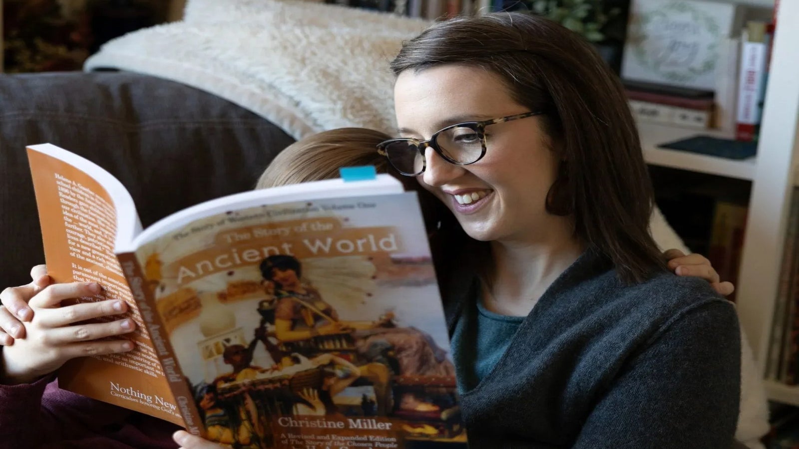 A woman and a child sit together on a couch reading a book titled The Story of the Ancient World by Christine Miller.