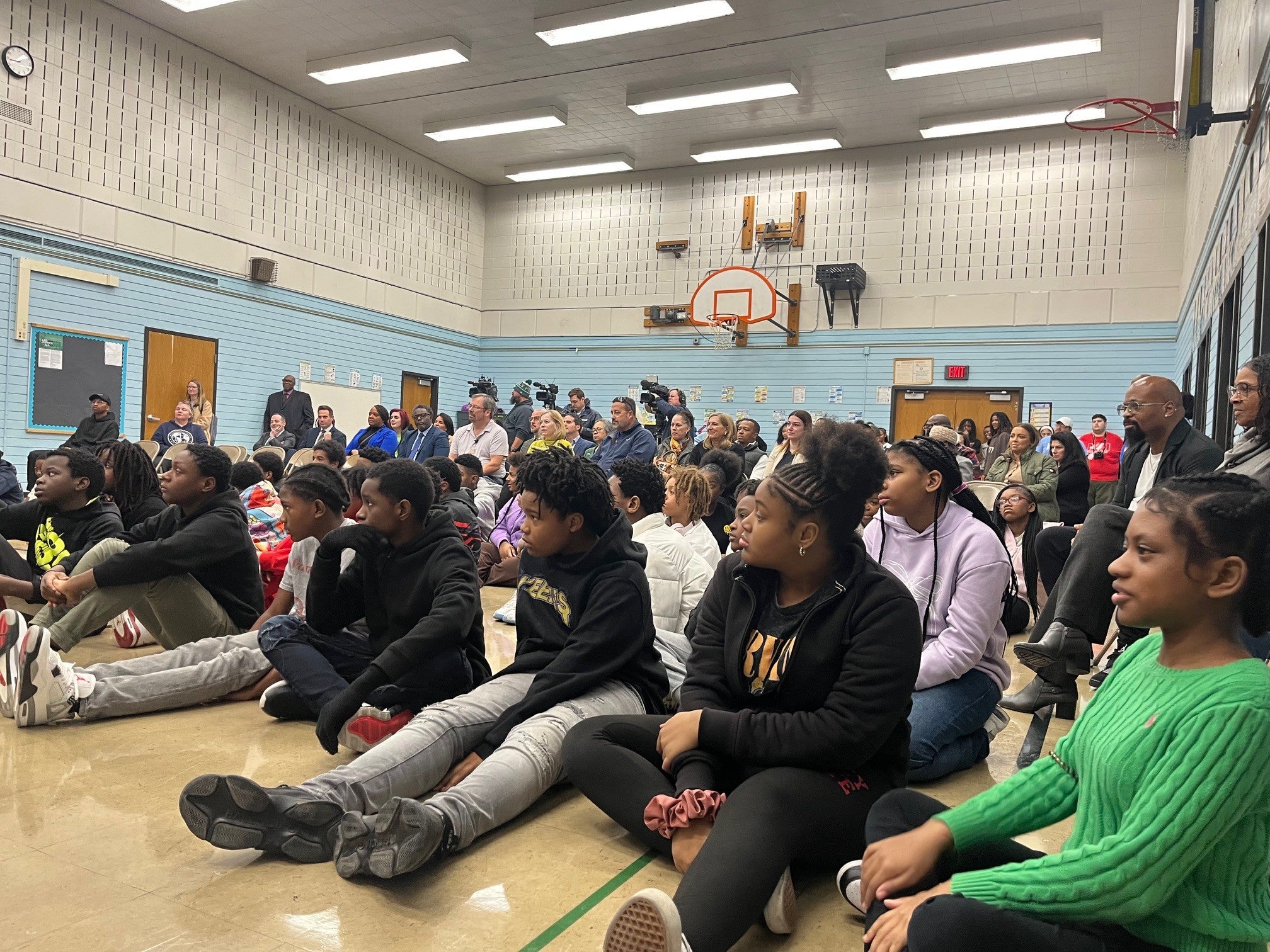 A group of students sit on the gym floor while adults sit on chairs behind them, all facing forward during an event in a school gymnasium.