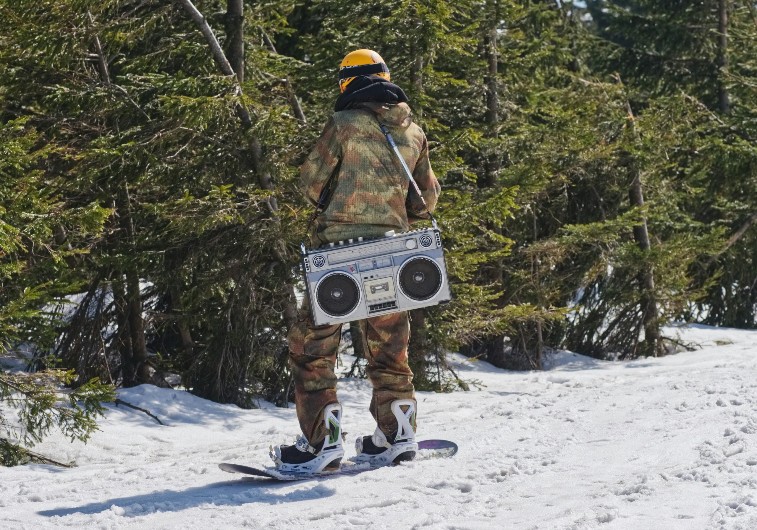 A person in camouflage clothing and a yellow helmet snowboards on a snowy slope while carrying a large boombox.