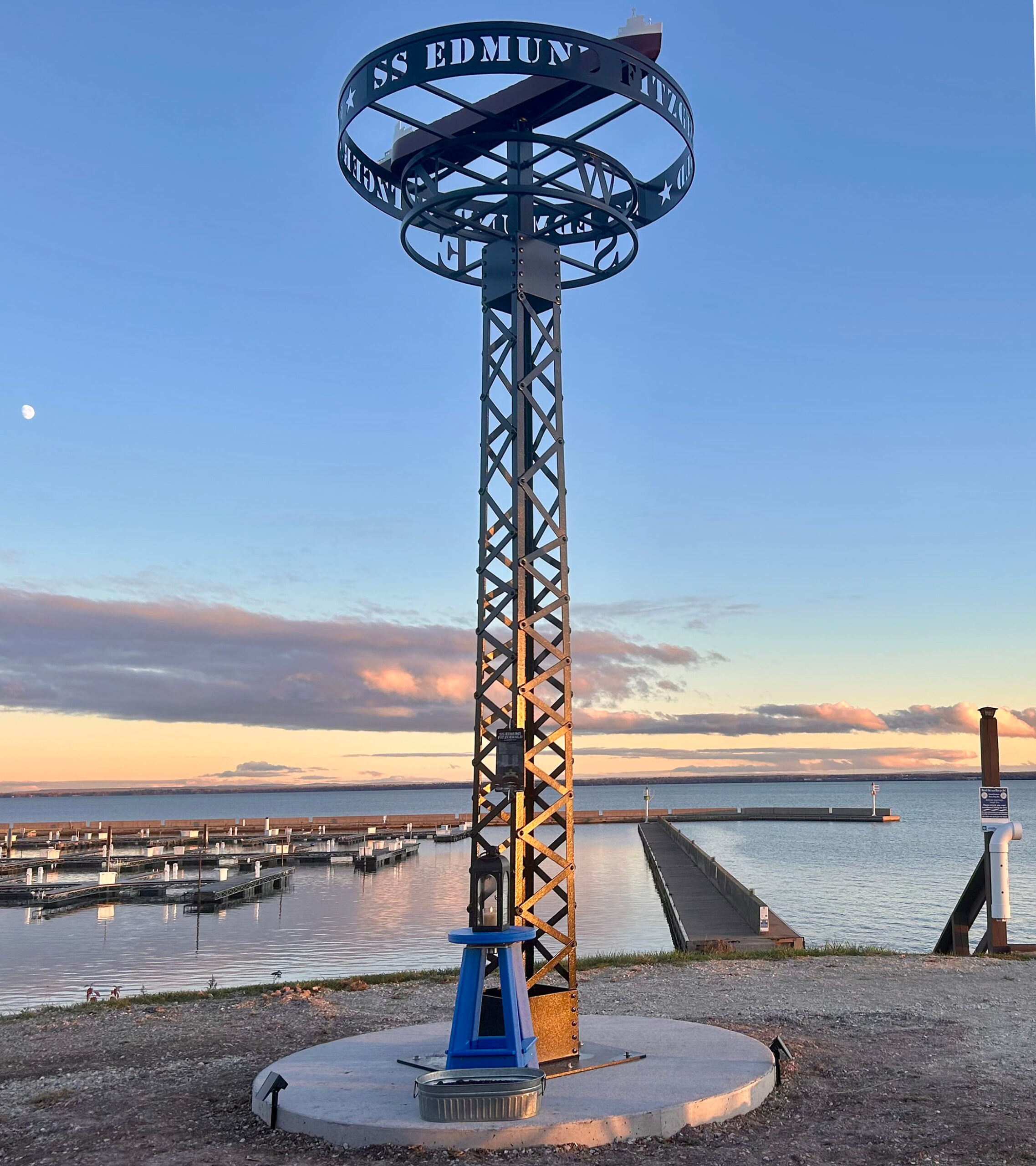 Metal memorial structure labeled SS Edmund Fitzgerald stands near a harbor with docks and water at sunset.