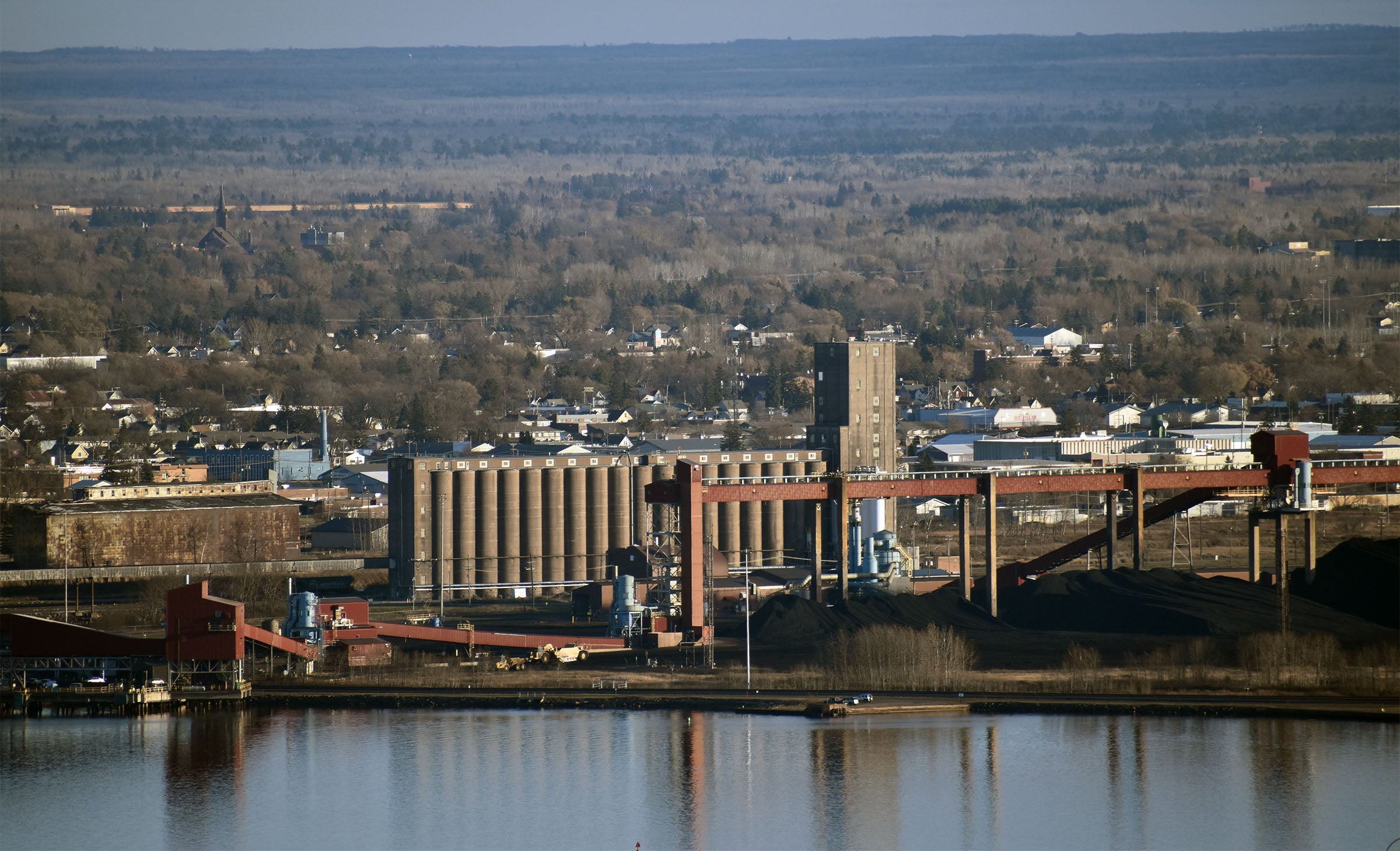Industrial complex with silos and conveyors near a waterfront, with a residential area and forested landscape in the background.