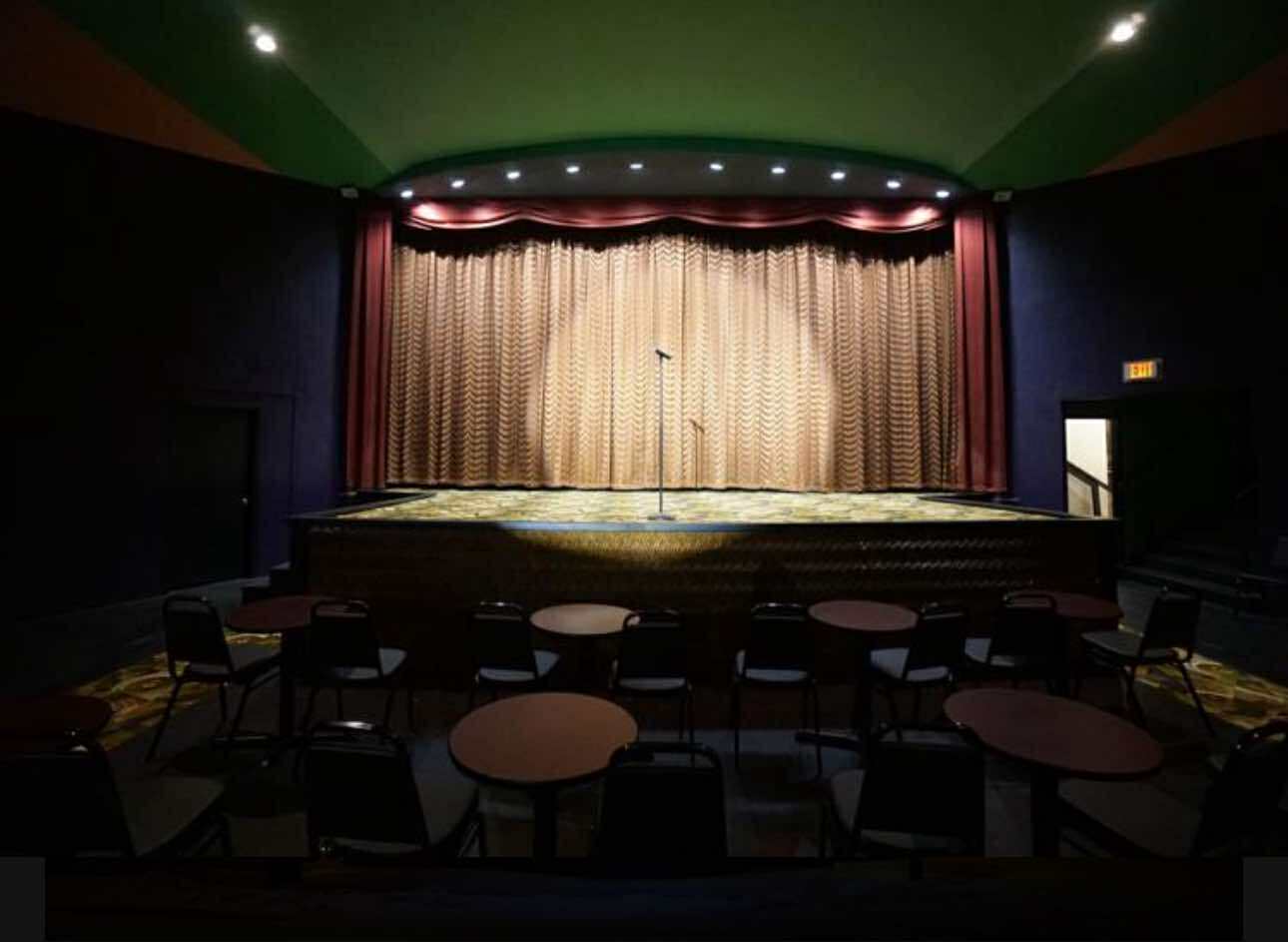 Empty stage with a single microphone in front of a closed curtain, facing rows of tables and chairs in a dimly lit theater.