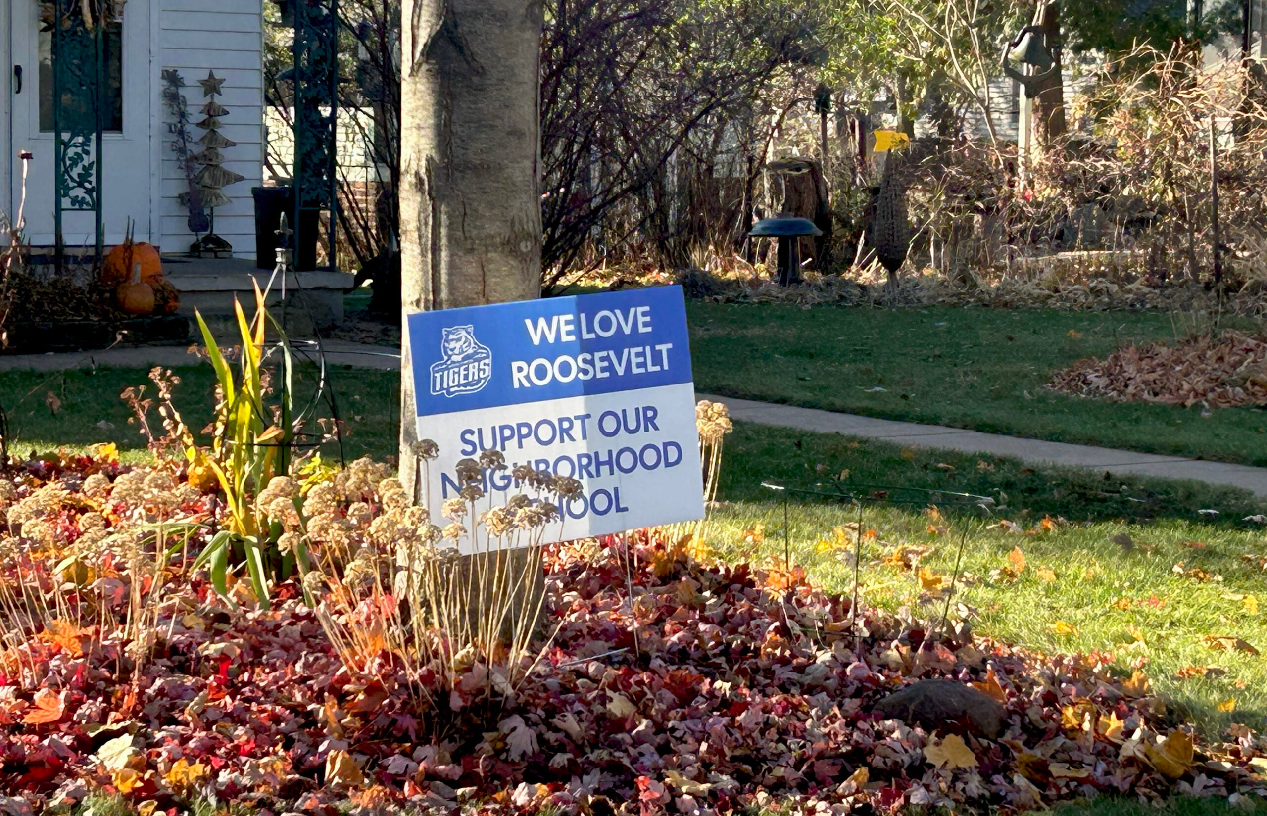 A sign in a yard reads We Love Roosevelt. Support Our Neighborhood School with fallen leaves and a house in the background.