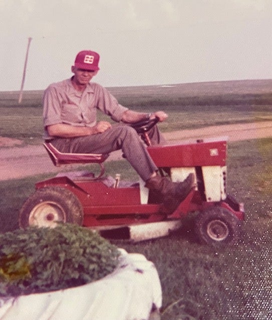A man wearing a red cap and matching shirt sits on a red riding lawn mower in a grassy field with a dirt path in the background.