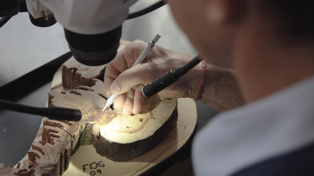 A person uses precision tools to carve or examine a wooden sample under a microscope, focusing on detailed work.