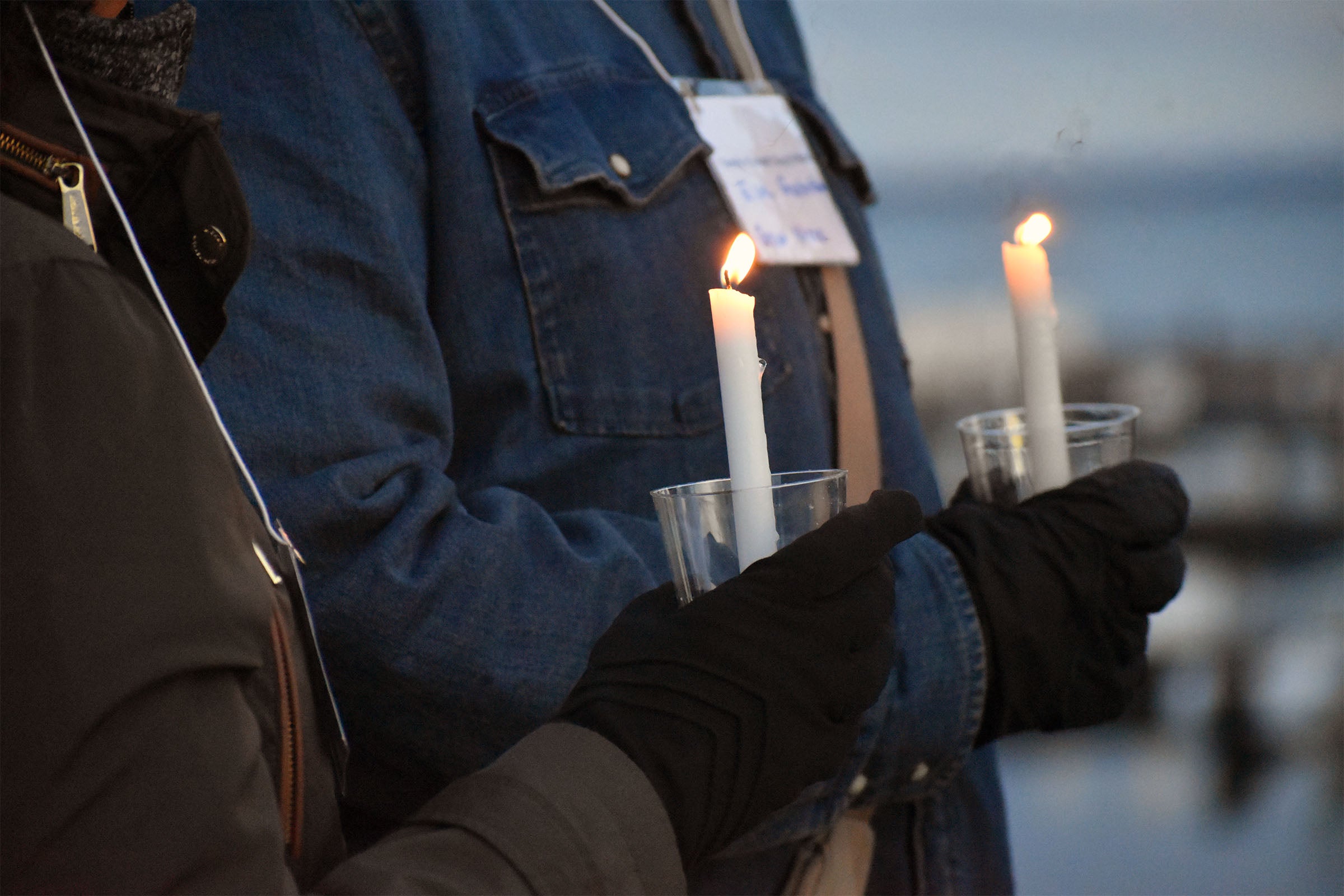 Two people wearing gloves and holding lit white candles in clear plastic cups, standing side by side at an outdoor event.