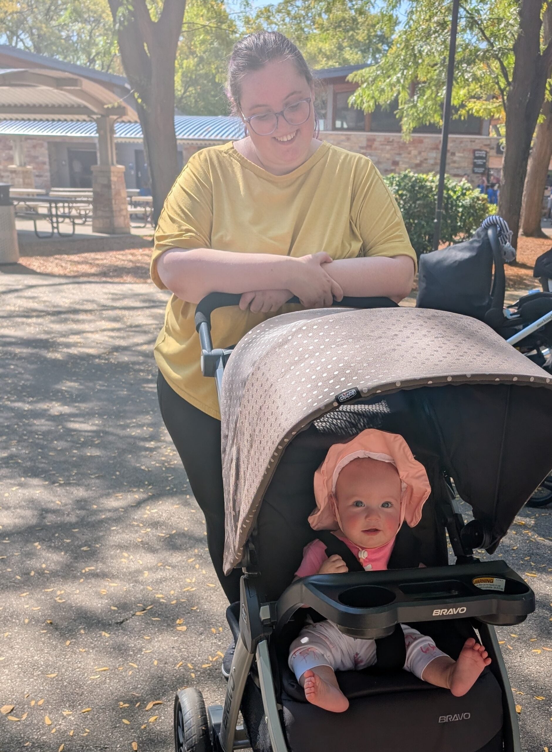 An adult stands behind a stroller, smiling at a baby who is sitting inside and wearing a pink hat. They are outdoors on a sunny day with trees and a pavilion in the background.
