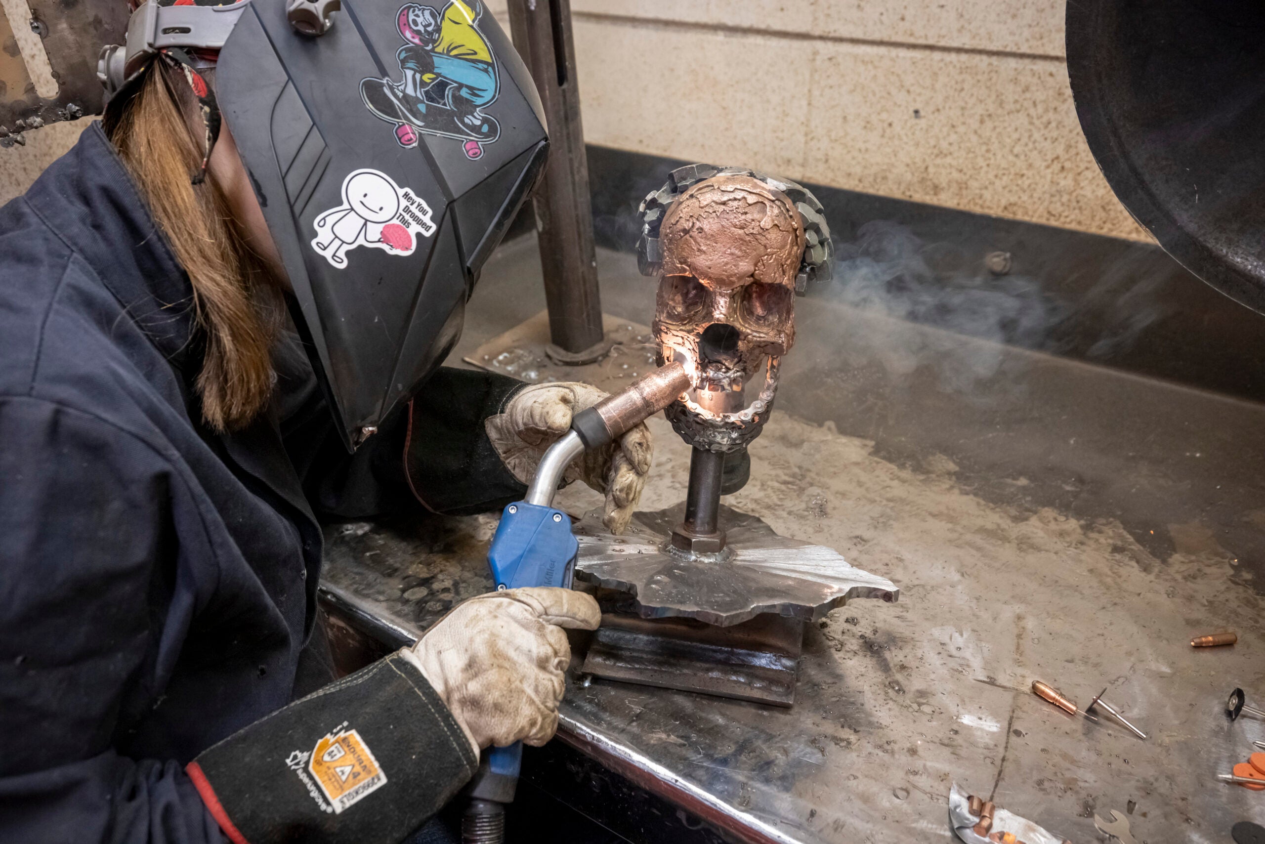 A person wearing protective gear uses a welding torch on a metal sculpture of a skull in a workshop.