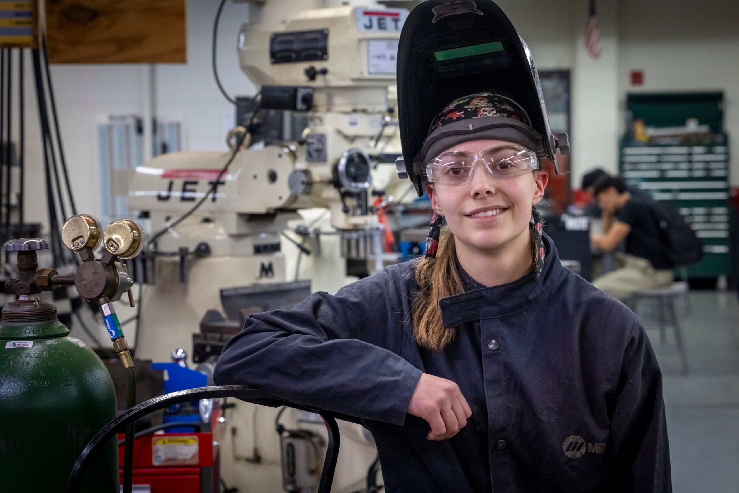 A person in safety gear stands in a workshop next to welding equipment, smiling at the camera. Industrial machinery and tools are visible in the background.