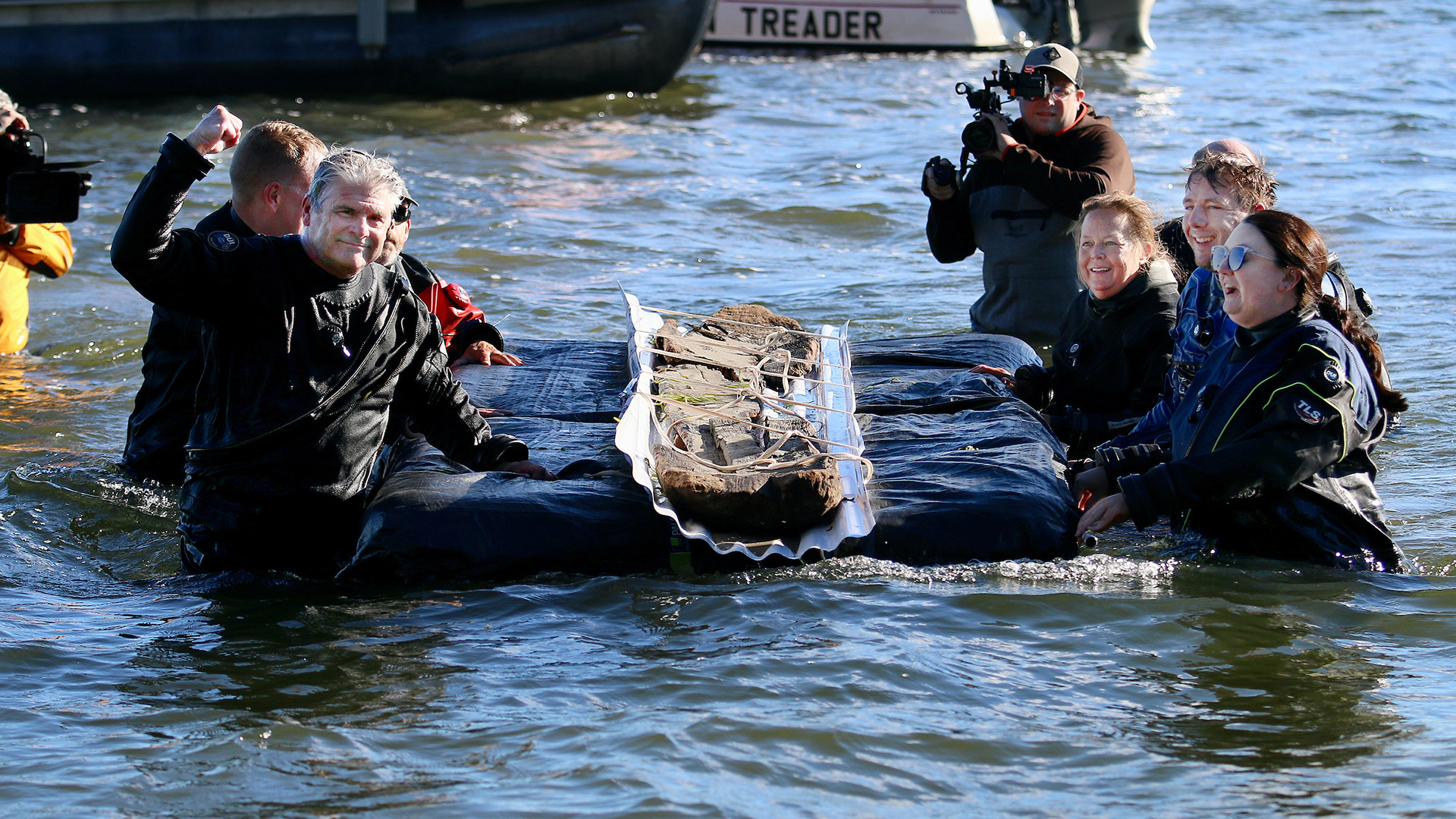 A group of people in water guide and support a large, ancient wooden artifact secured on a floating platform, with cameras recording the event.