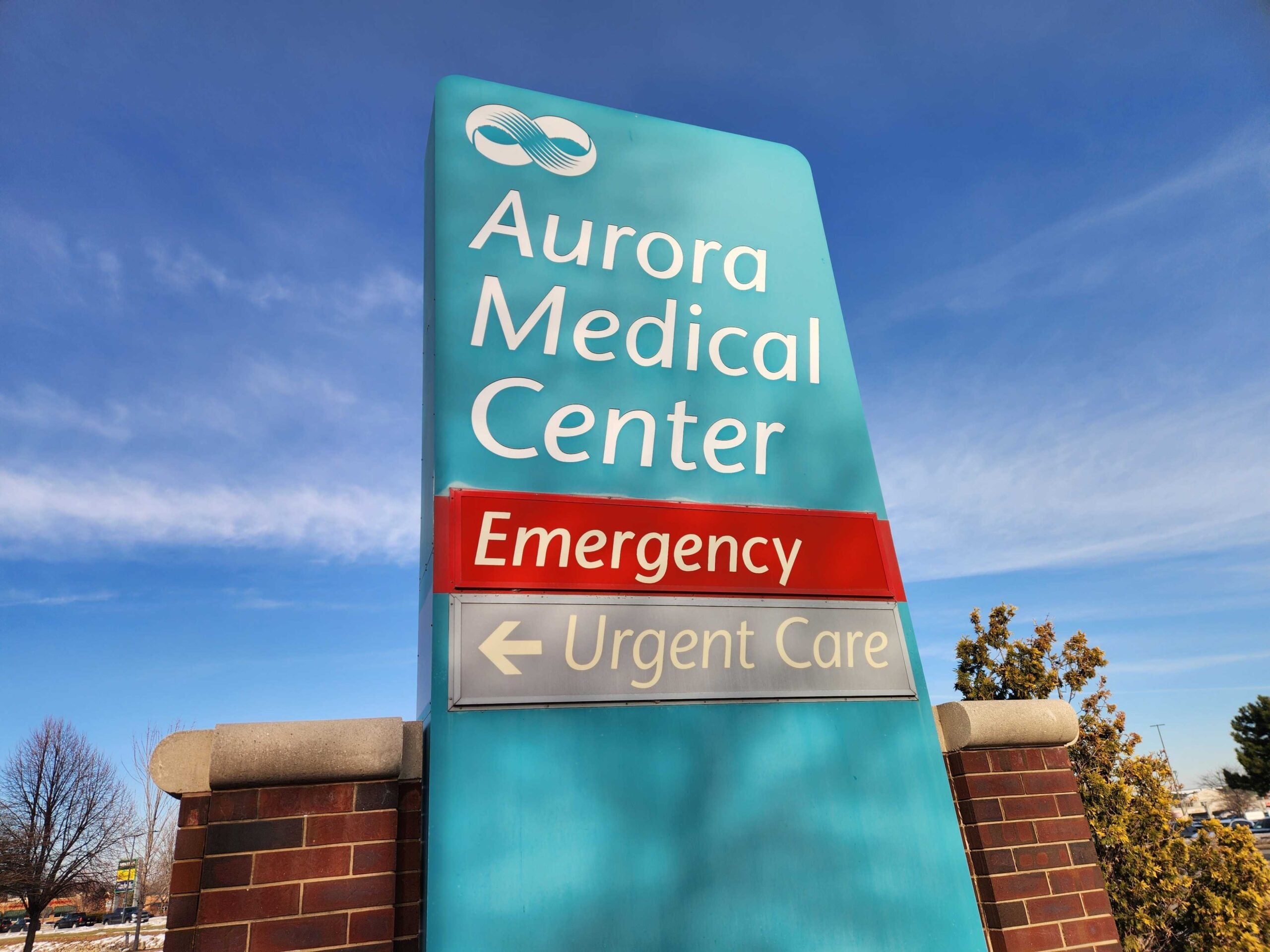 Sign for Aurora Medical Center with labels for Emergency in red and Urgent Care below, set against a blue sky background.
