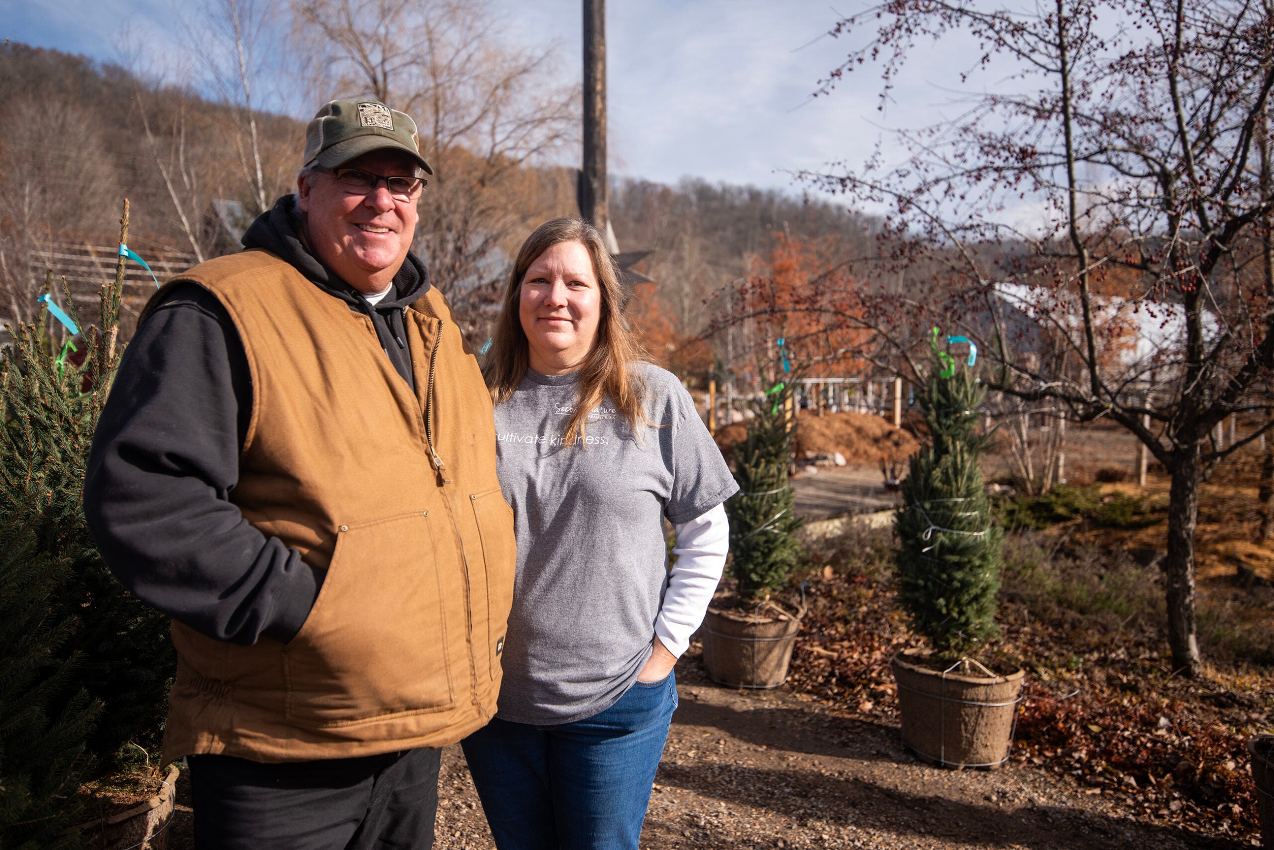 A man and woman stand outdoors in a tree nursery on a sunny day, with potted evergreen trees and leafless trees around them.