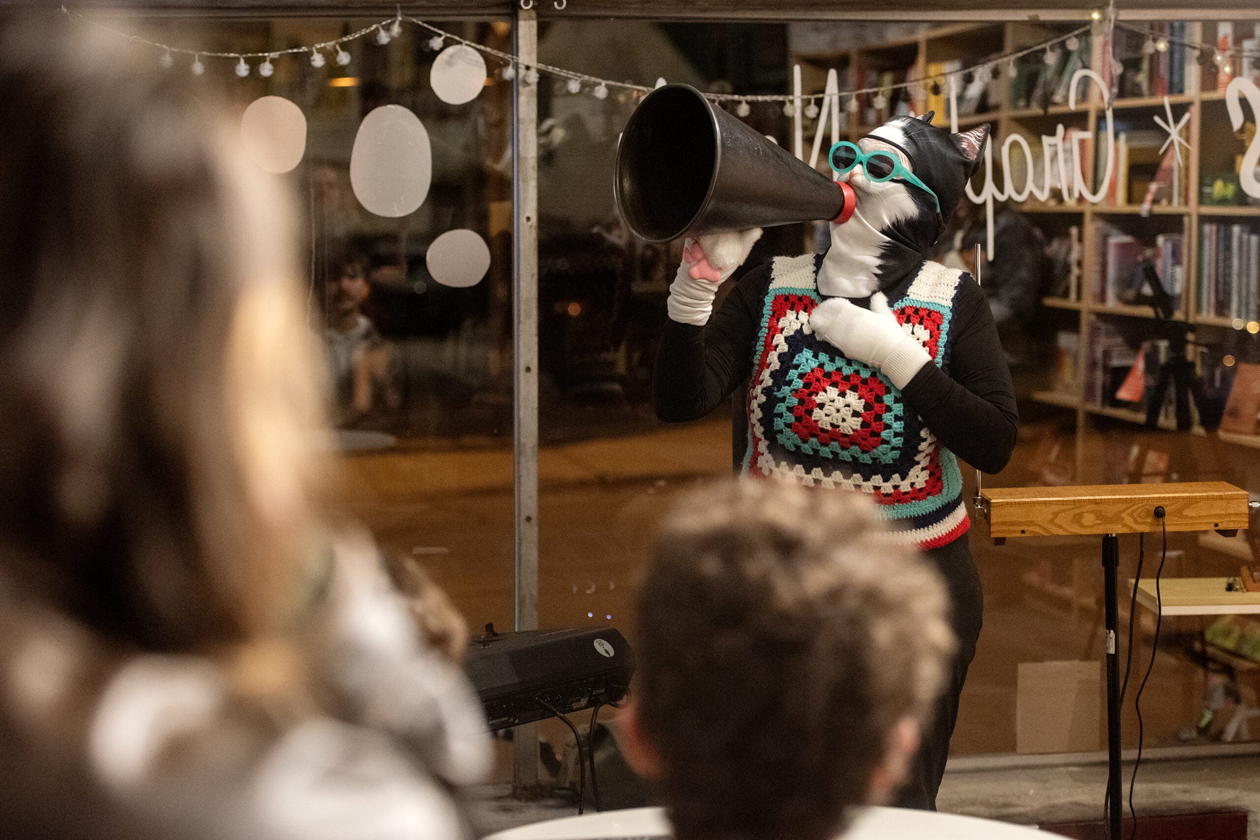 A person in a cat mask, crochet vest, and teal glasses uses a megaphone to address an audience in a room decorated with string lights and bookshelves.