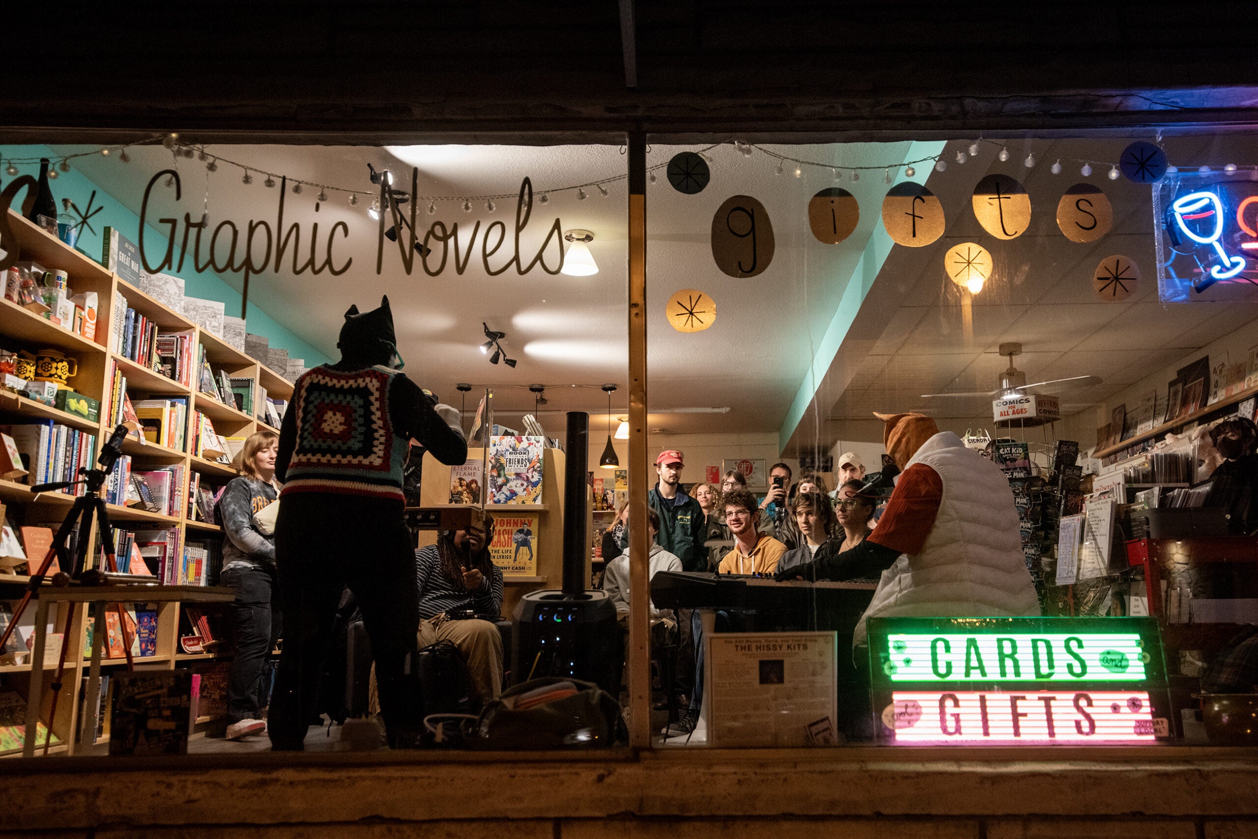People gather inside a bookstore decorated with signs for graphic novels and gifts, while someone in a cat hat speaks to the group. Shelves of books and cards are visible.