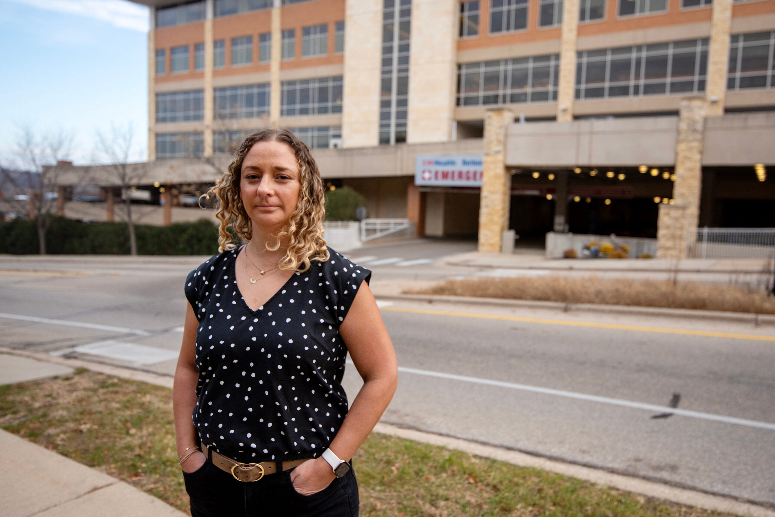A woman with curly hair, wearing a black and white polka dot shirt, stands on a sidewalk in front of a multi-story building and an emergency entrance sign.