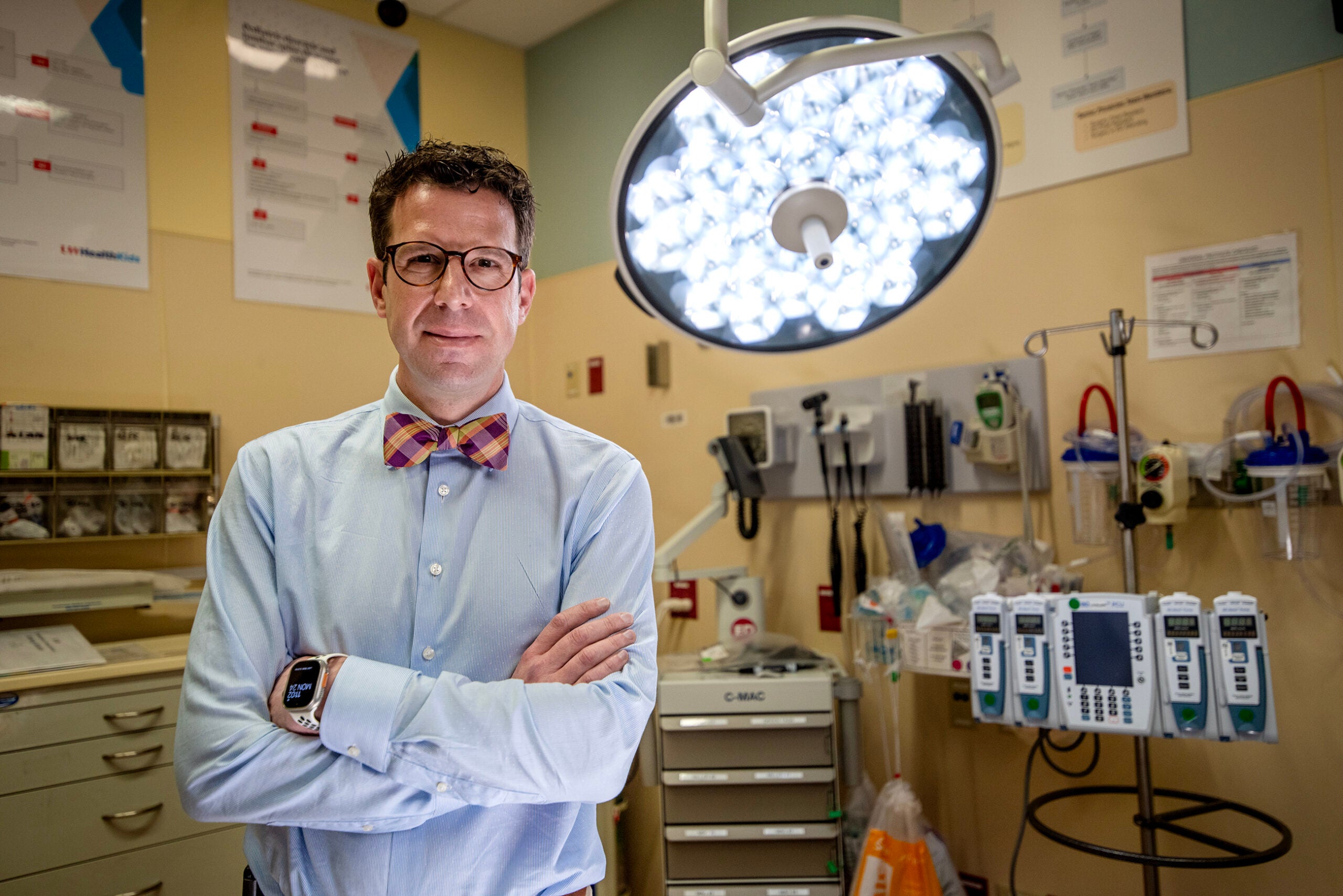 A man in a dress shirt and bow tie stands with arms crossed in a hospital room, surrounded by medical equipment and supplies.