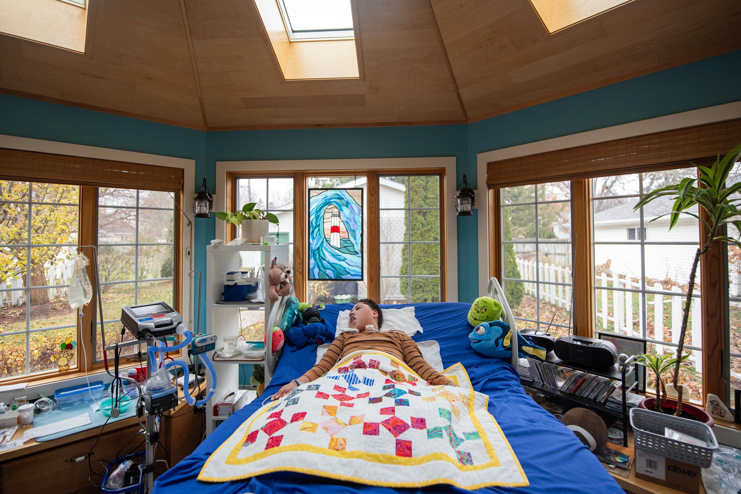 A person lies in a bed covered with a colorful quilt in a sunlit room with large windows, surrounded by medical equipment, plants, and a stained glass window above the bed.