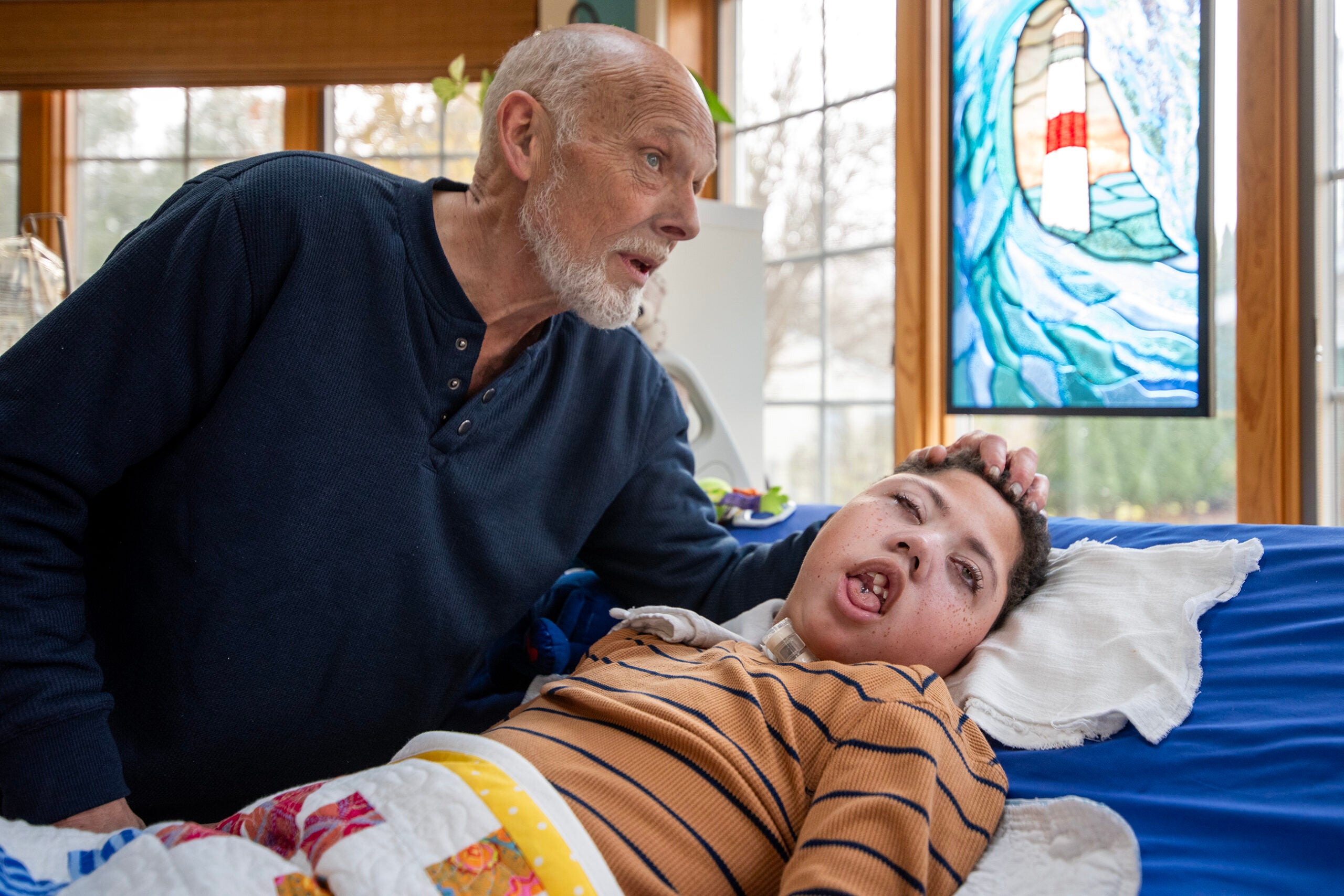 An older man gently holds the head of a young boy lying in bed, who appears to be unresponsive. A stained glass window is visible in the background.
