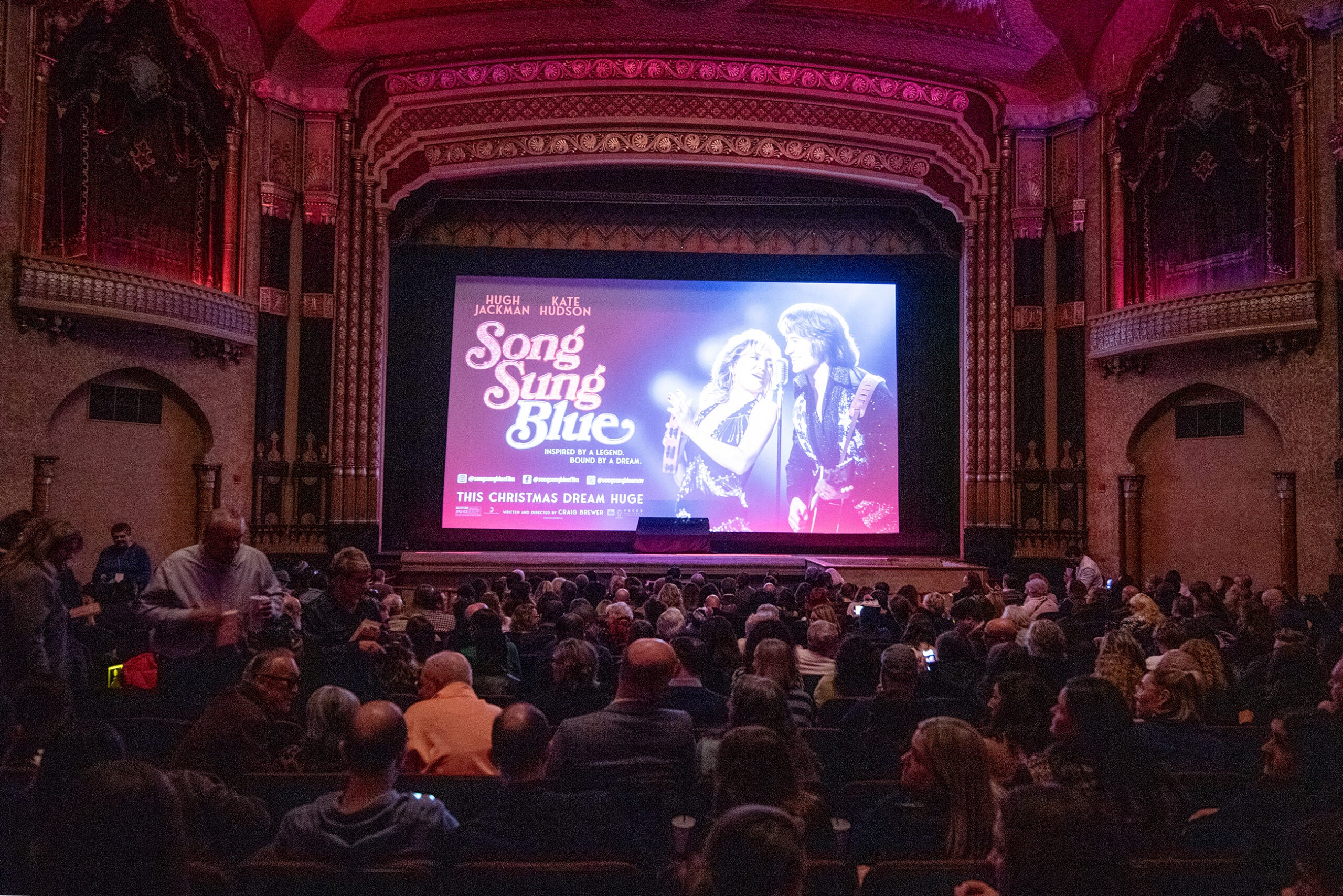 Audience seated in a historic theater with ornate decor, facing a large screen displaying the Song Sung Blue movie poster featuring two singers.