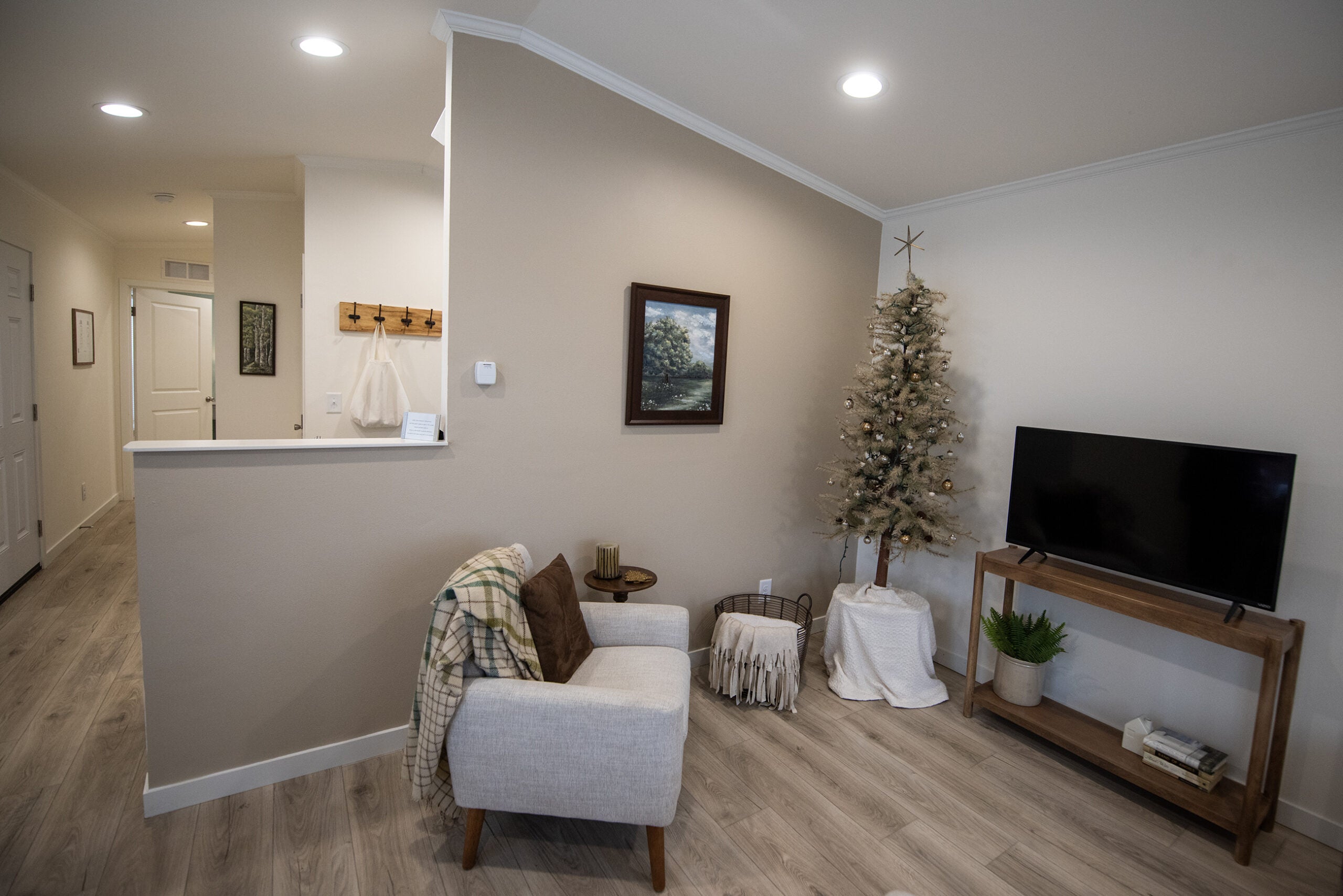 A small living room features a gray armchair, a TV on a wooden stand, a minimalist Christmas tree, and neutral decor with light wood floors and beige walls.