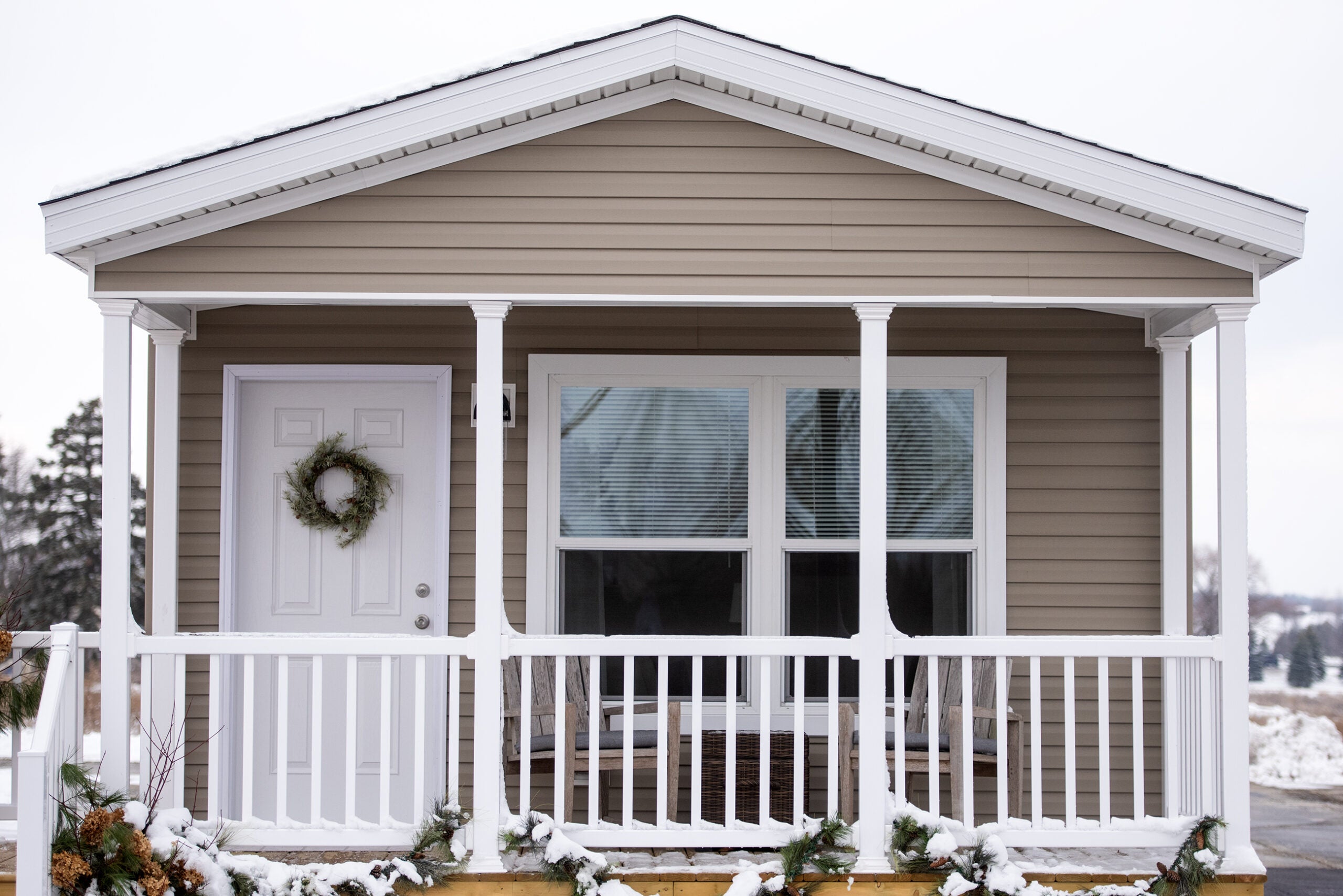A small beige house with white trim, a front porch, and a wreath on the door. Snow is on the ground and the porch railing is decorated with garland.