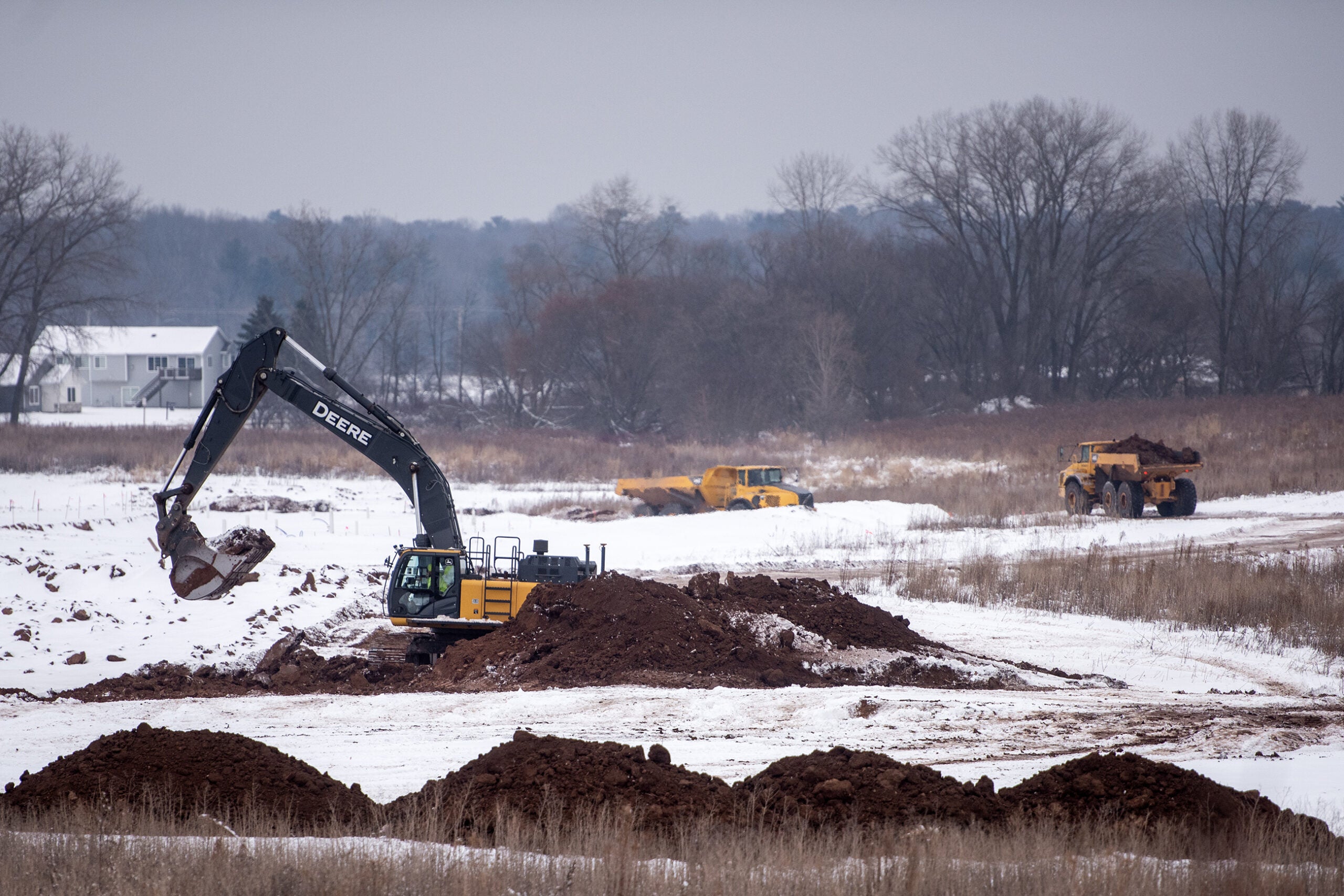 An excavator and two dump trucks move soil on a snow-covered construction site in a rural area with bare trees and a house in the background.