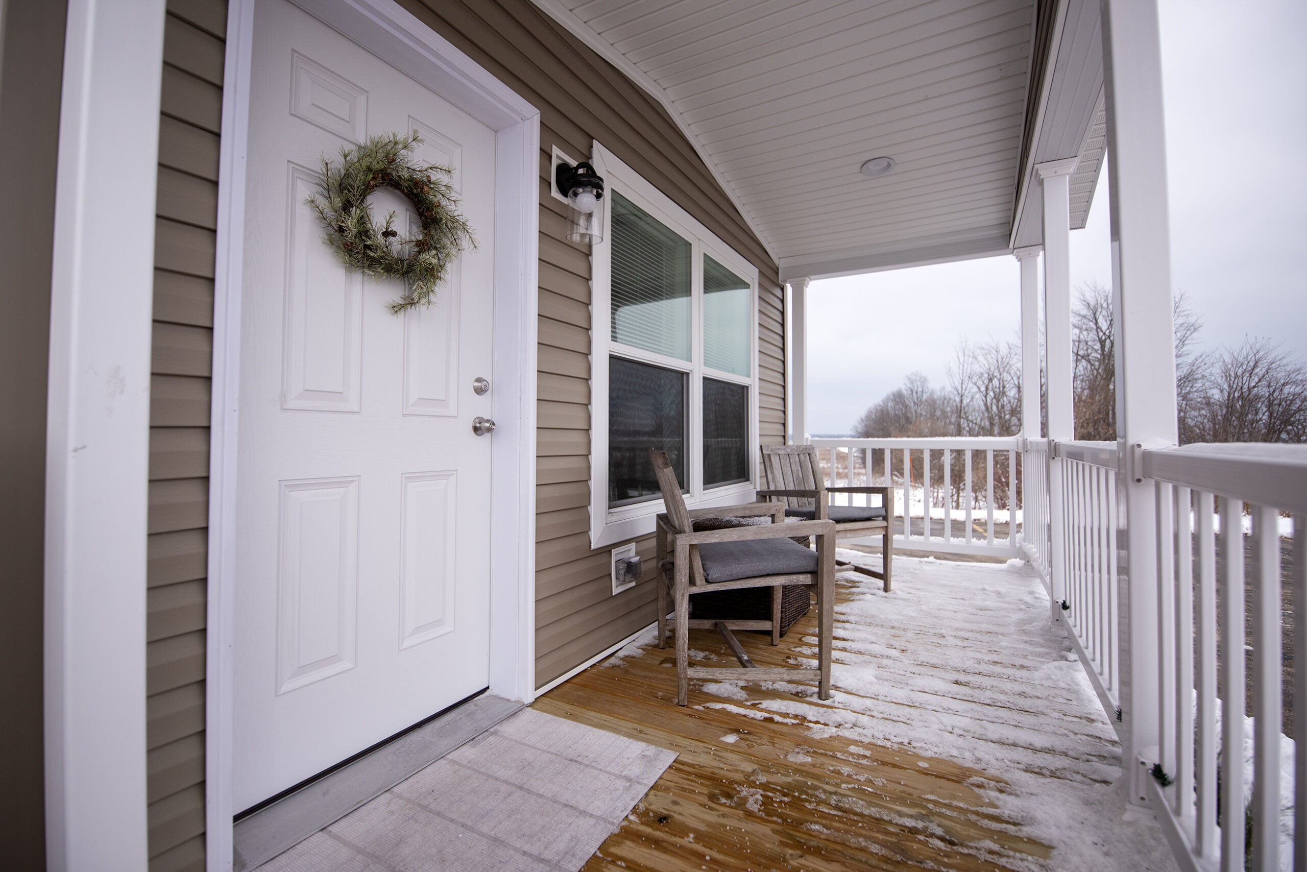 A snowy front porch with wooden chairs, a small table, white railing, and a white door decorated with a wreath.