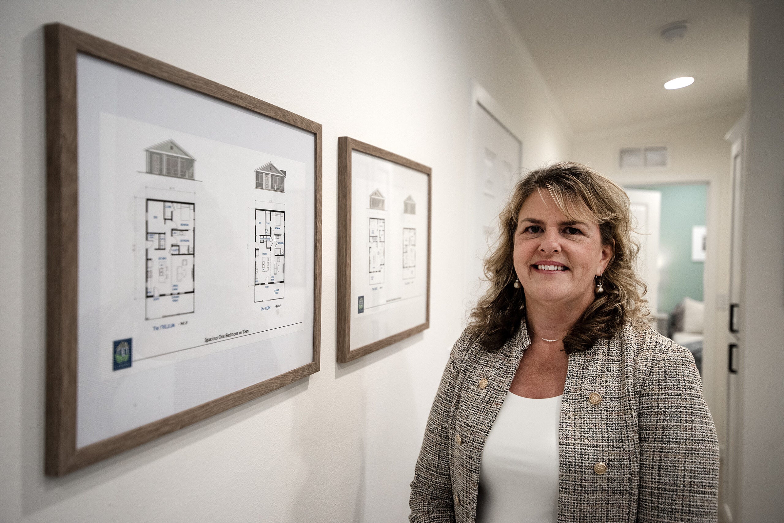 A woman stands indoors next to framed architectural floor plans mounted on a wall.