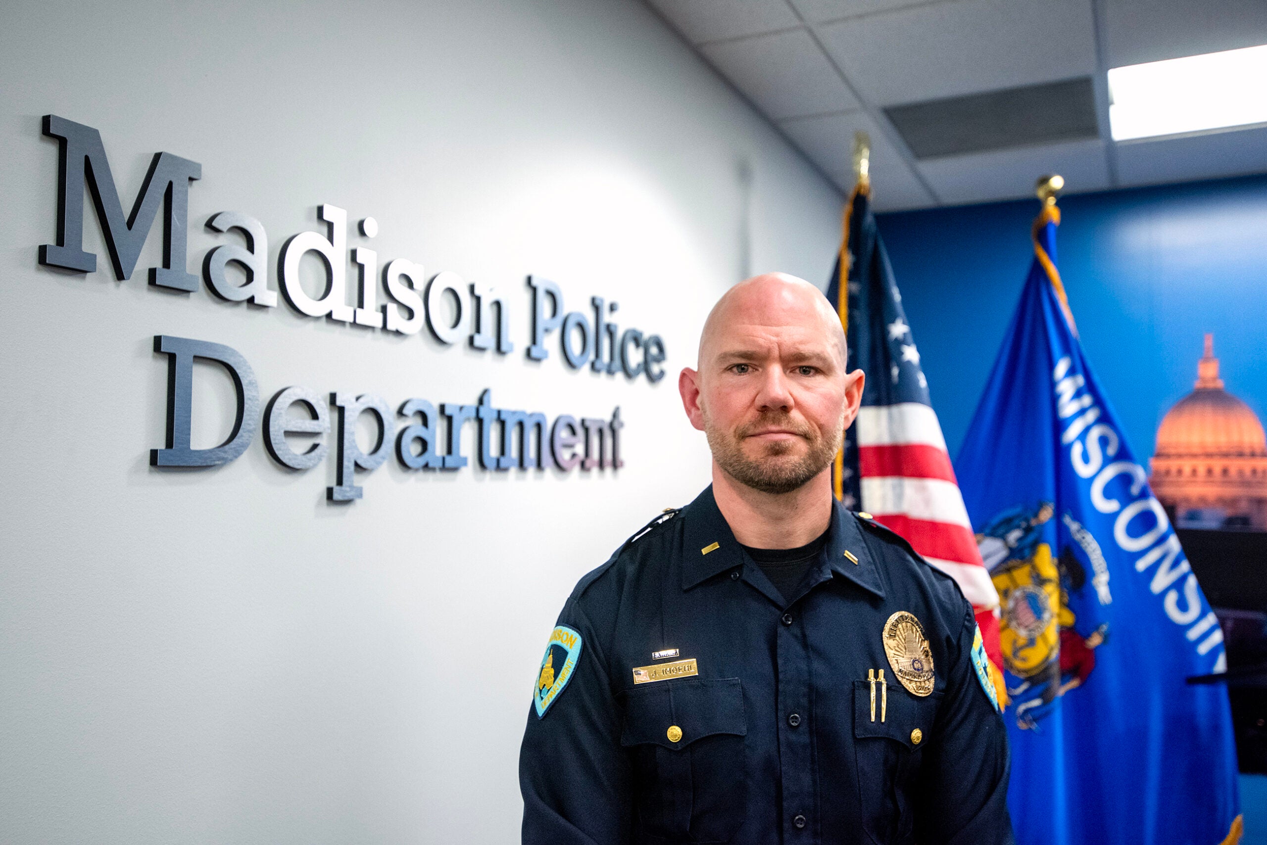 A police officer in uniform stands in front of a wall sign reading Madison Police Department with U.S. and Wisconsin flags in the background.