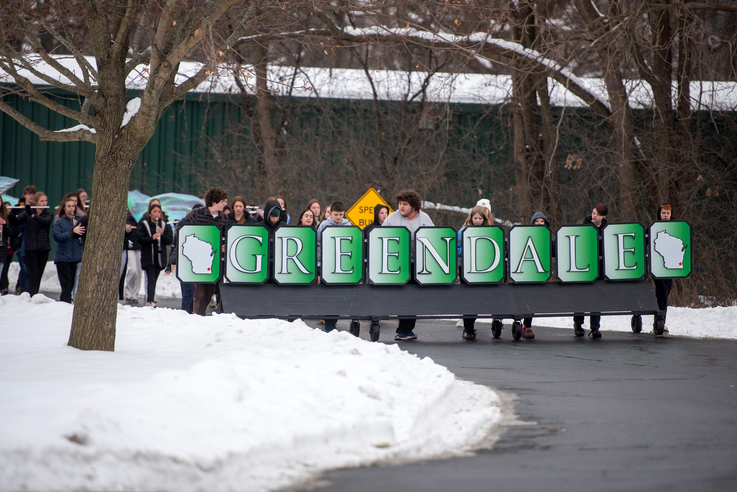A group of people walk on a snowy road, holding large signs that spell out Greendale with silhouettes of Wisconsin on each letter.
