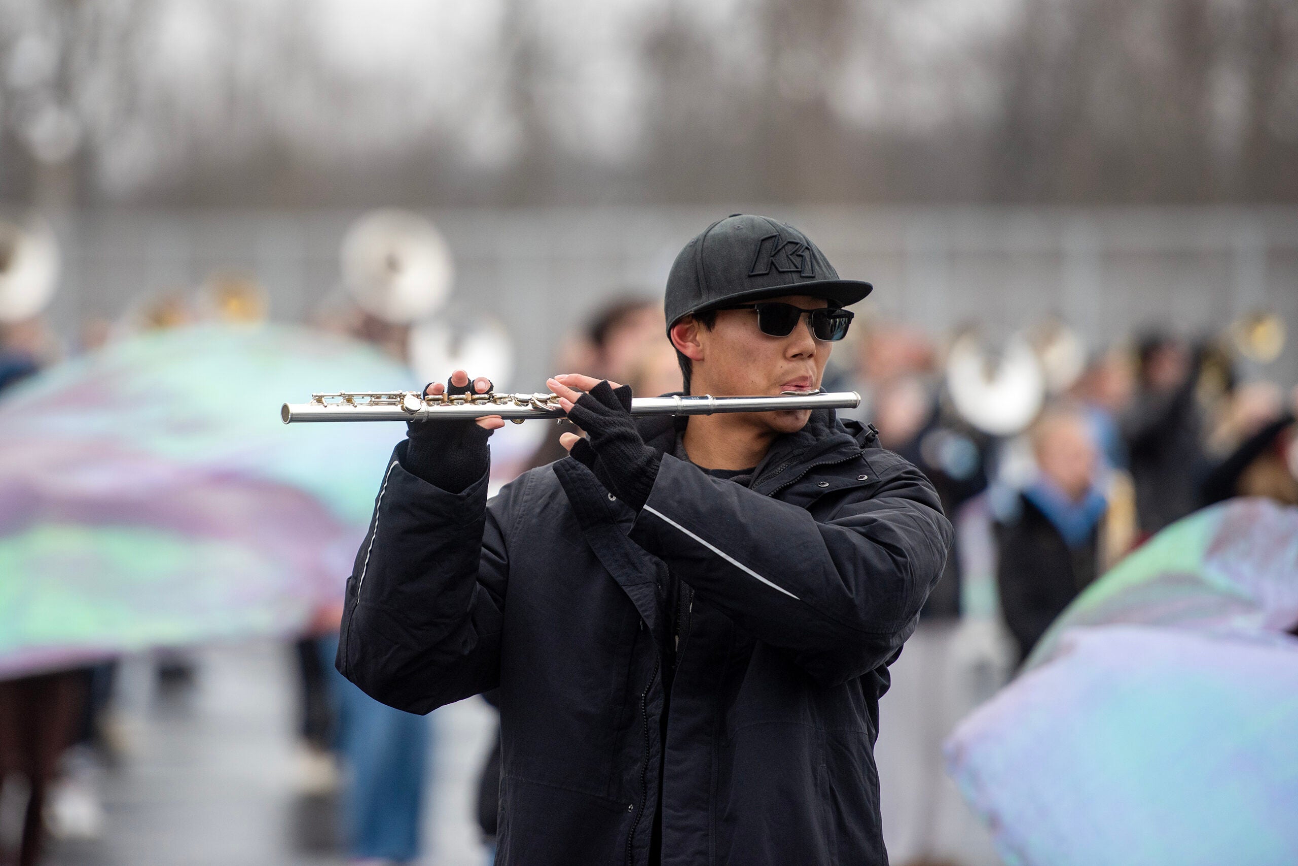 A person wearing sunglasses and a black cap plays the flute outdoors, with other band members and colorful flags visible in the blurred background.