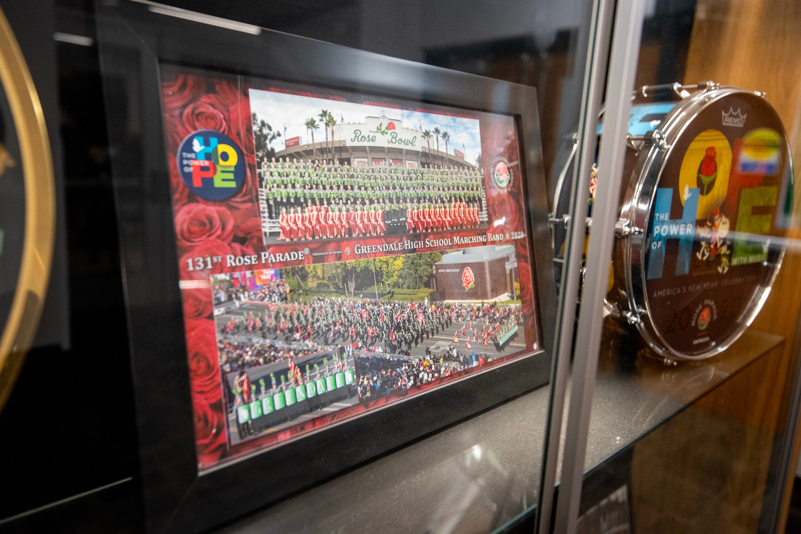 A framed photo collage of the Greendale High School Marching Band at the 131st Rose Parade is displayed in a glass case next to a decorated drum.