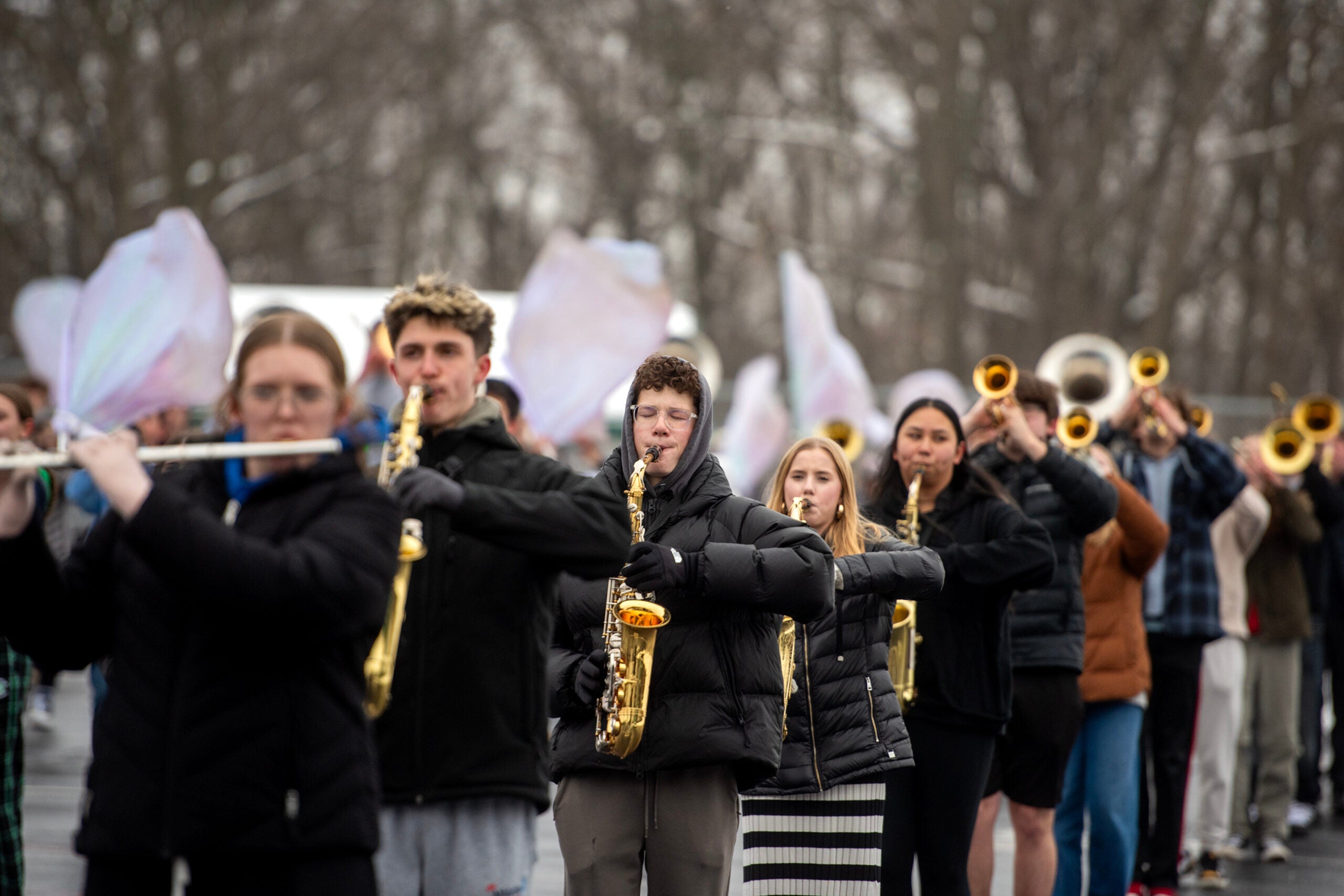 A marching band practices outdoors in cold weather, with members playing saxophones, flutes, and trumpets, while others hold large white flags in the background.