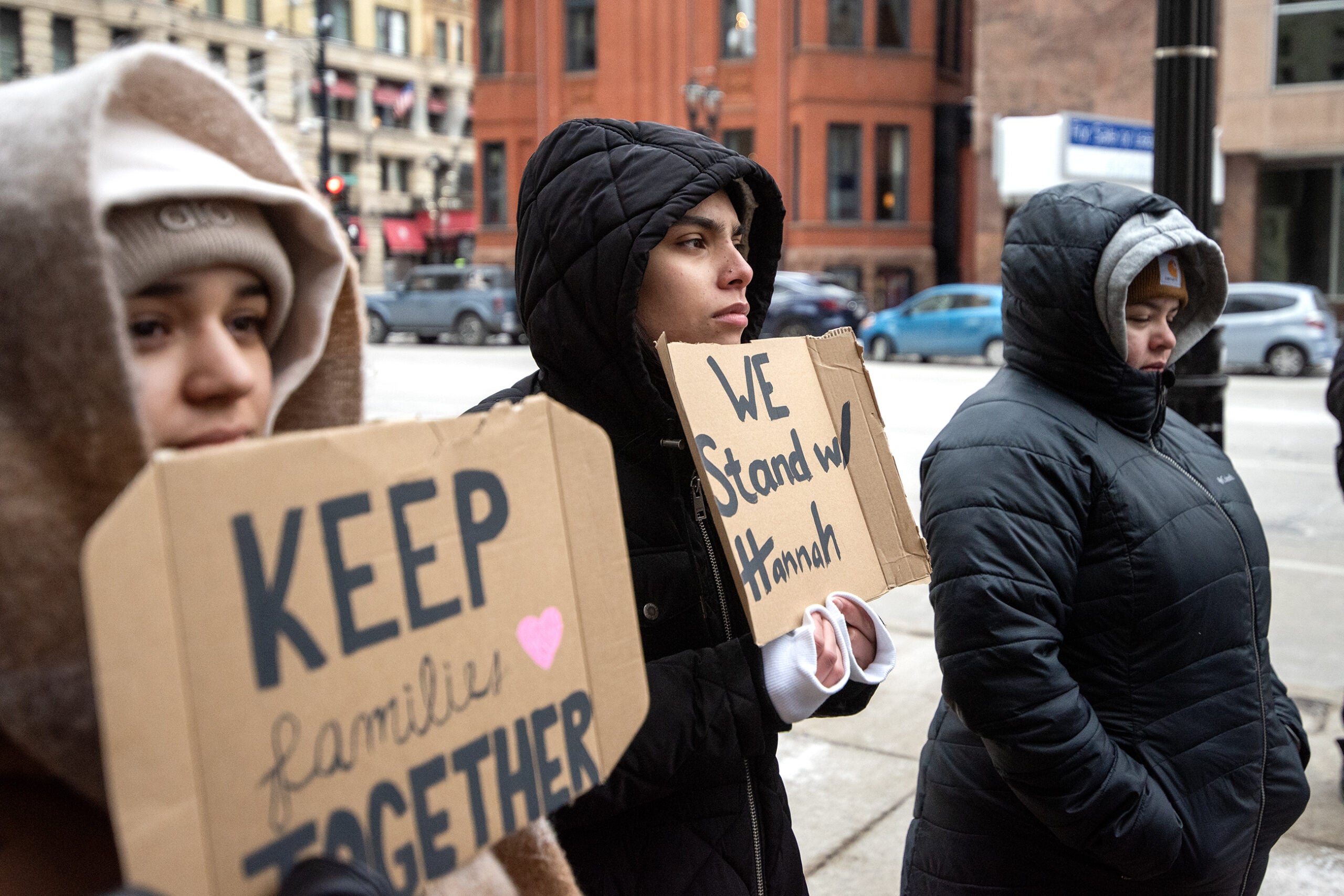 Three people in winter coats hold cardboard signs reading KEEP Families TOGETHER and WE Stand w/ Hannah while standing on a city sidewalk.