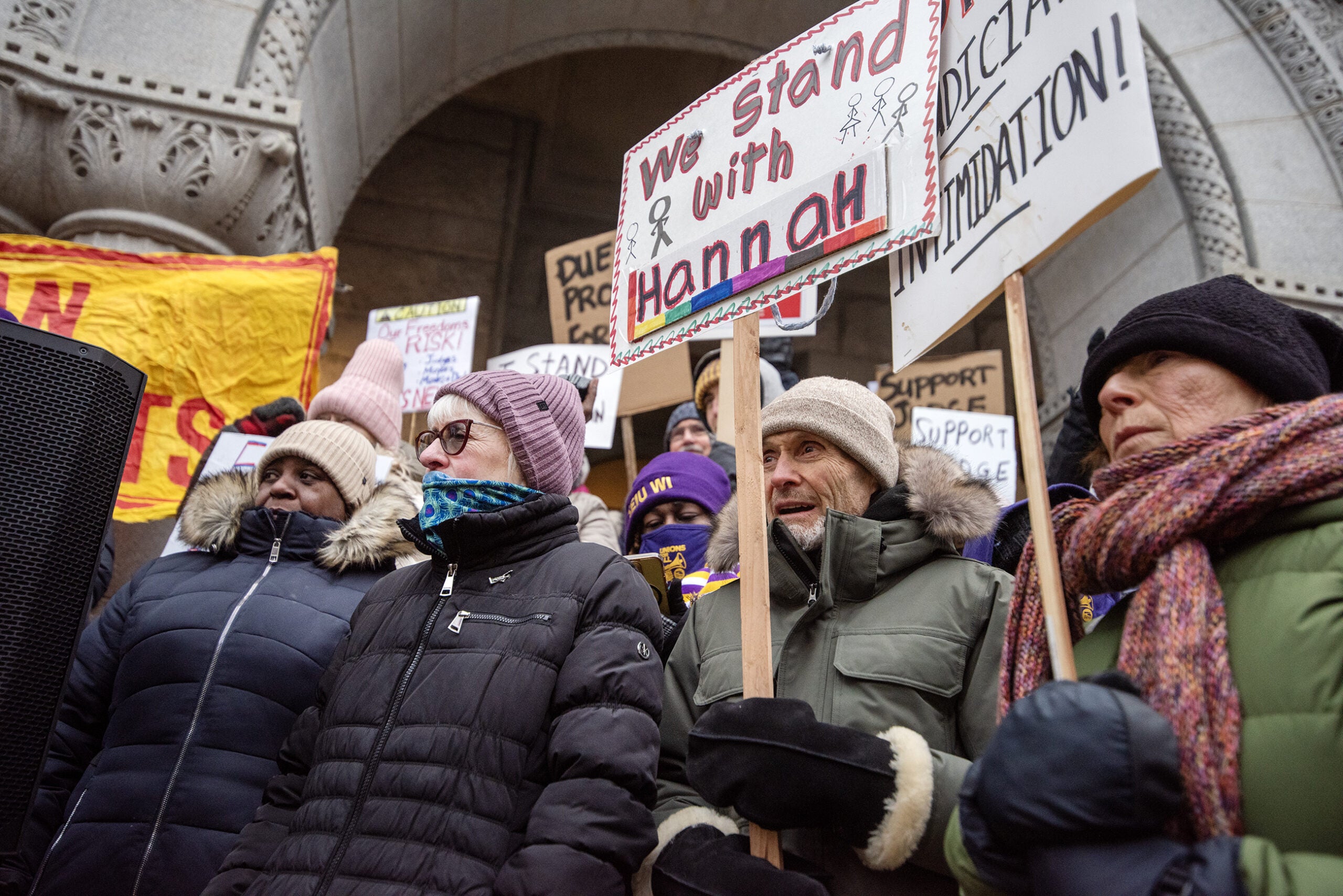 A group of people in winter clothing hold protest signs, including one that says We stand with Hannah, outside a building with arched stonework.