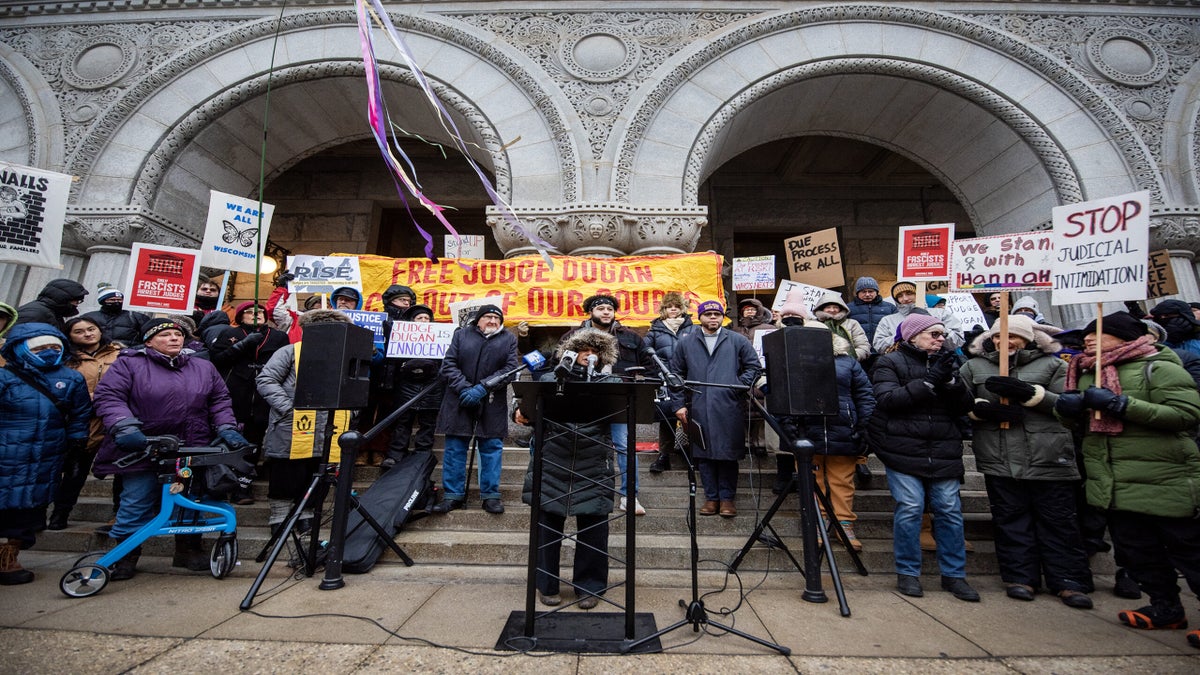A group of people stand on courthouse steps holding signs and banners at a protest, with a speaker at a podium in front of microphones.