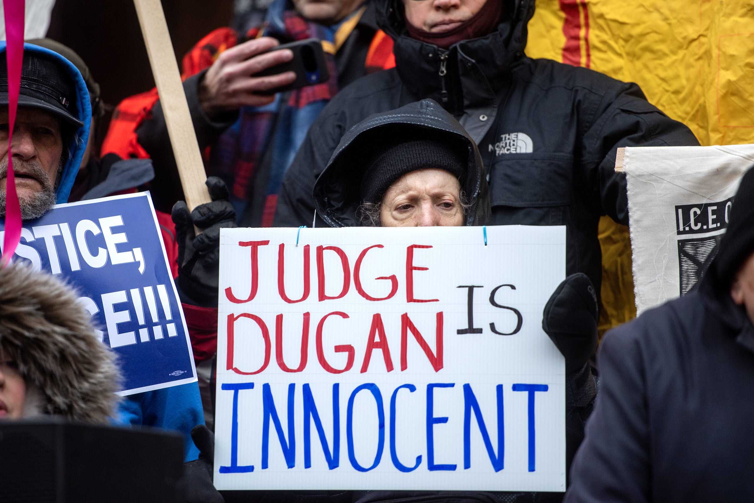 A person wearing a hooded jacket holds a sign that reads JUDGE DUGAN IS INNOCENT at a public gathering or protest.