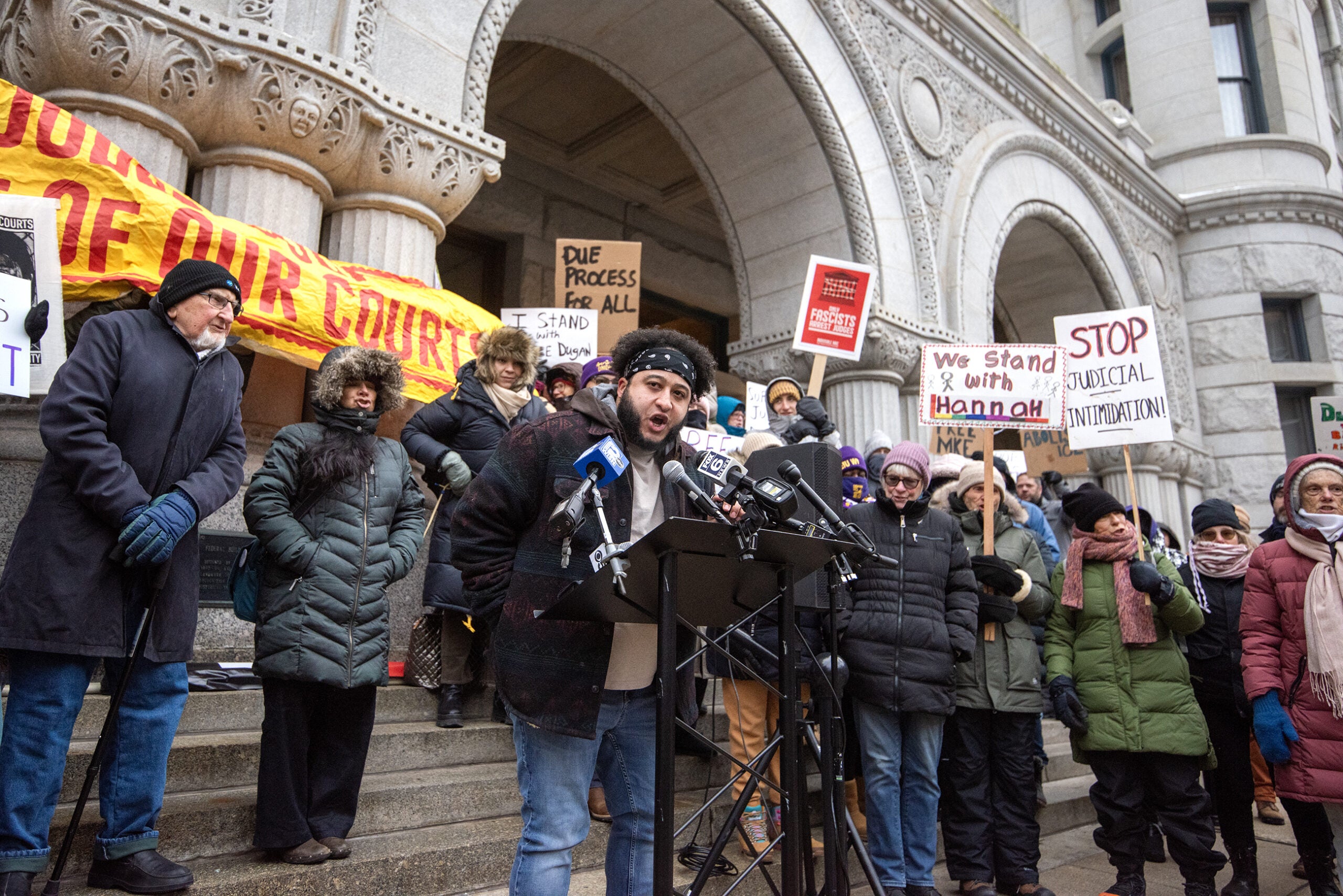 A group of people, bundled in winter clothing, hold protest signs and gather around a speaker at a podium outside a large stone building.