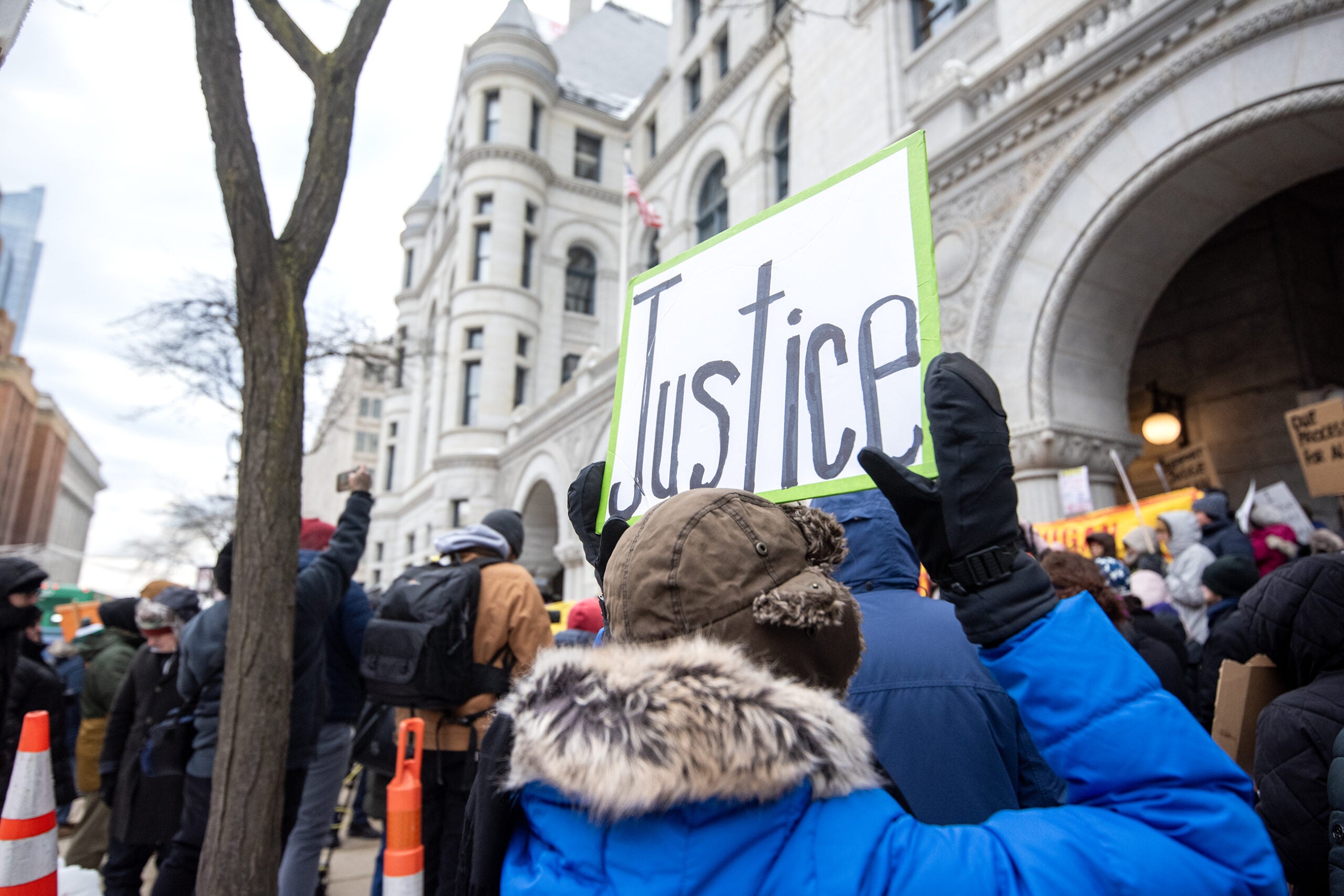 A person in winter clothing holds a sign reading Justice at a protest outside a large, stone building with an arched entrance.