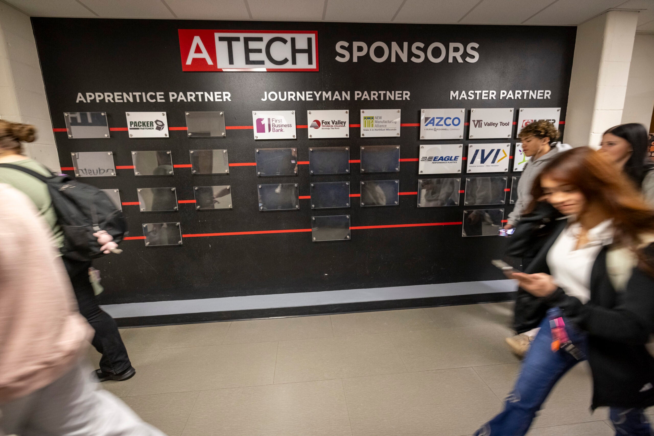 Students walk past a sponsor recognition wall labeled “ATECH Sponsors” with logos under Apprentice, Journeyman, and Master Partner categories.