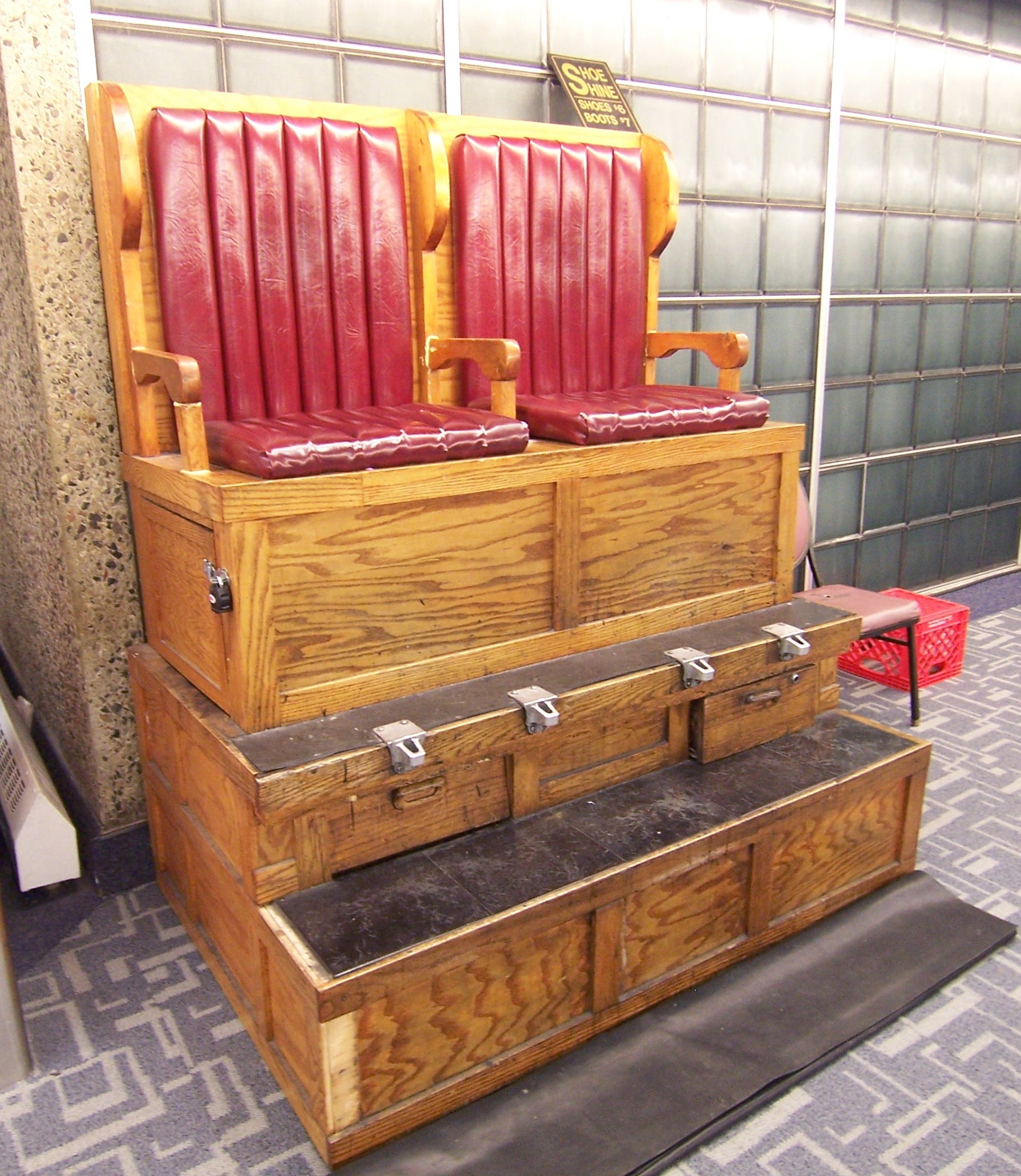 A wooden dual-seat shoe shine stand with red padded chairs and footrests, positioned on a carpeted floor in front of a glass block wall.