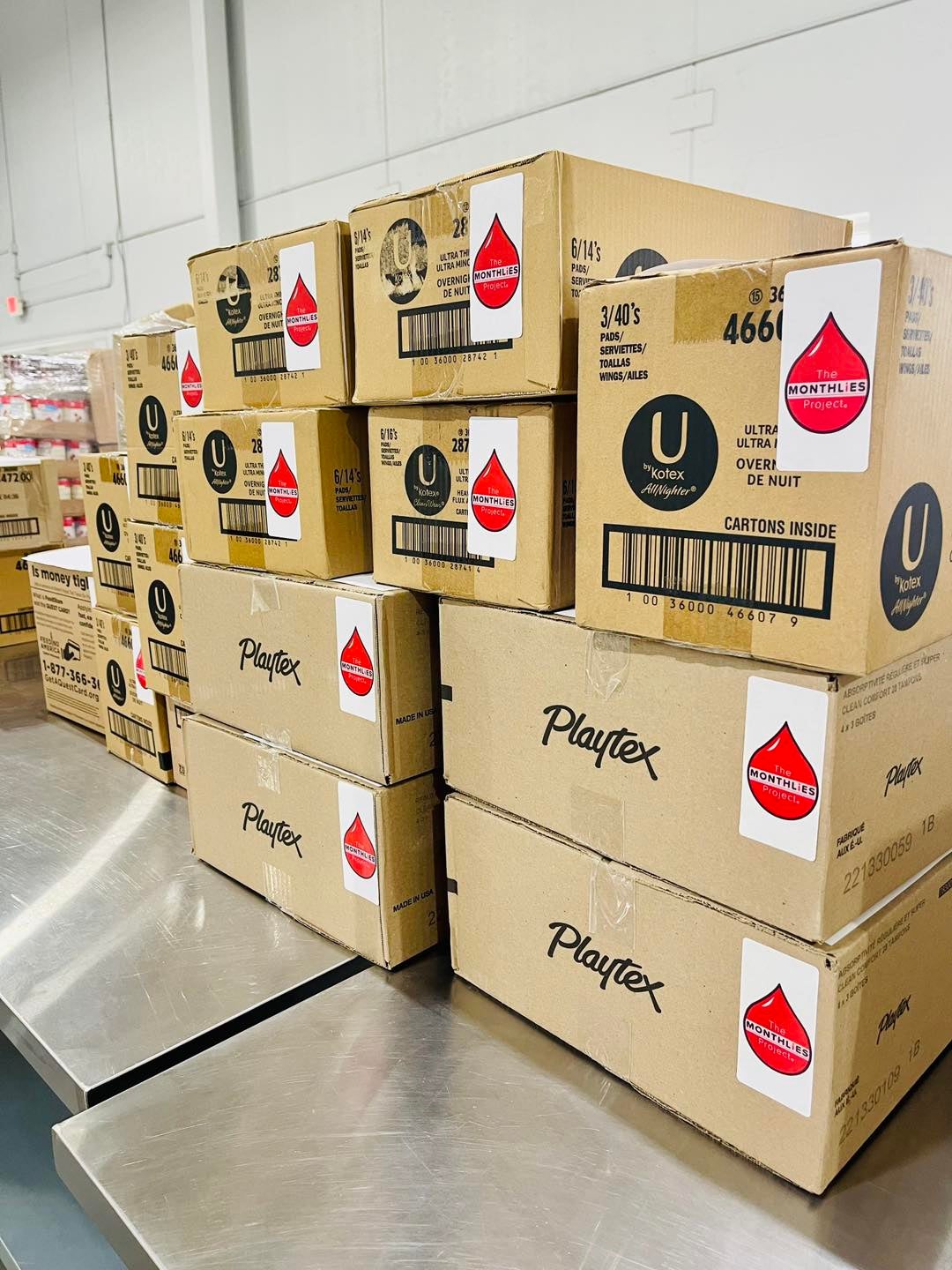 Stacks of cardboard boxes labeled Playtex and U by Kotex sit on a metal table in a warehouse or storage room.