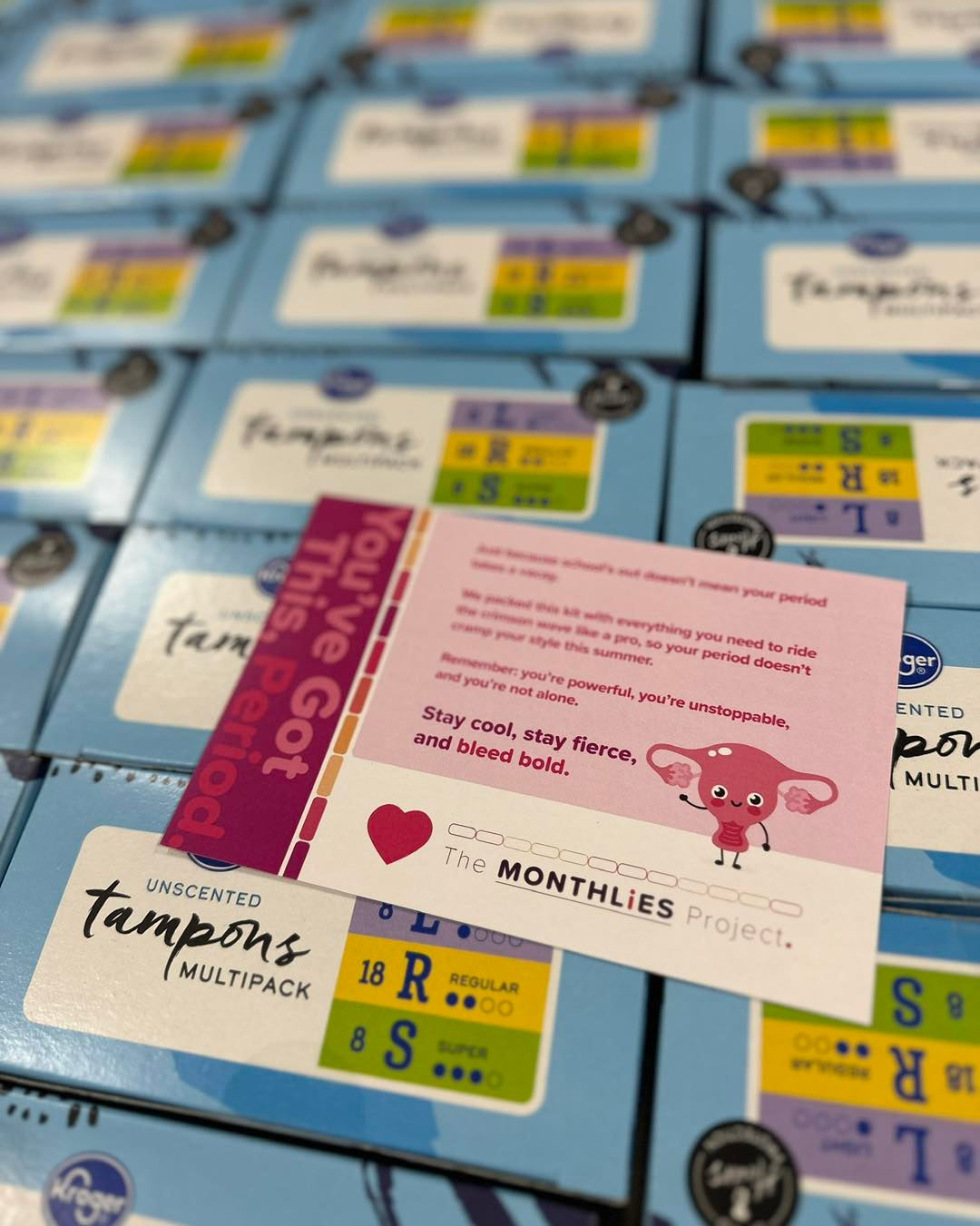 A card from The Monthlies Project sits on top of stacked boxes of unscented tampon multipacks, with information and encouragement about periods.