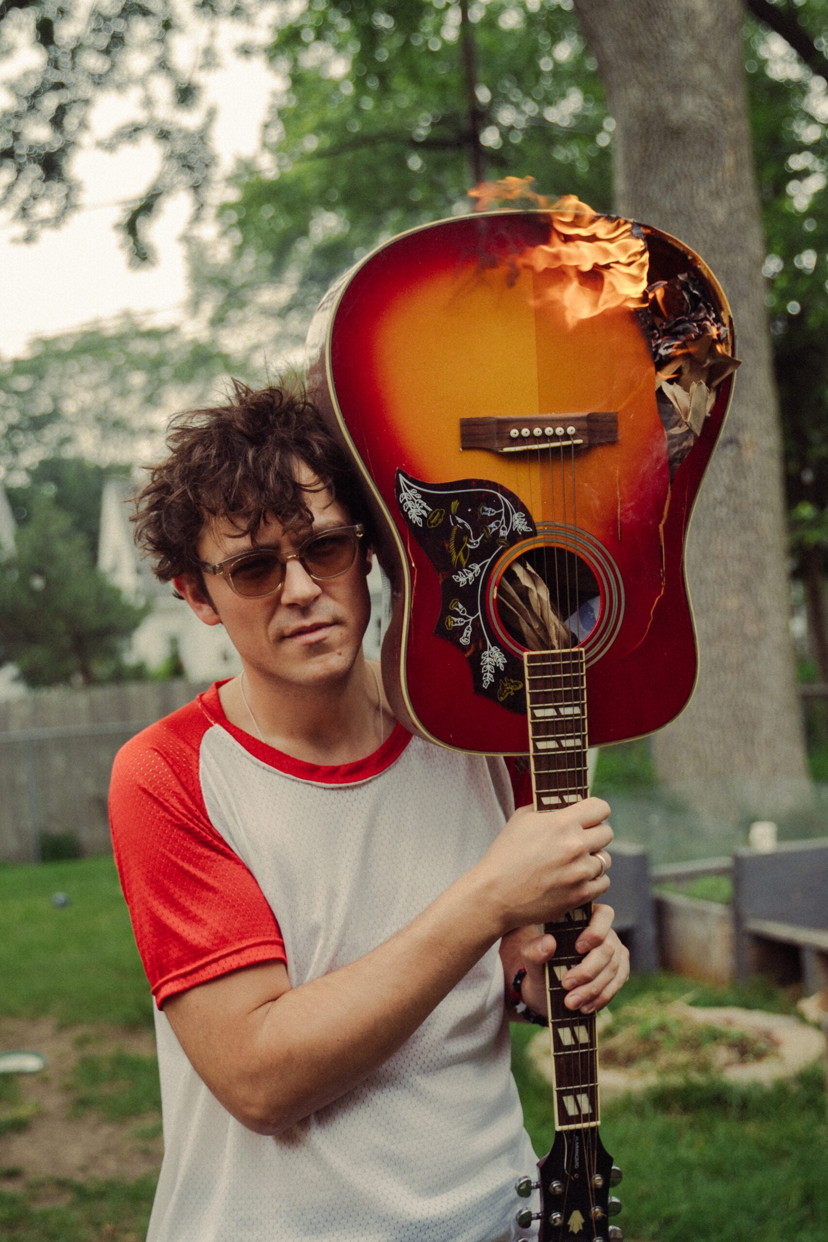 A person wearing sunglasses holds a burning acoustic guitar over their shoulder in a backyard setting.
